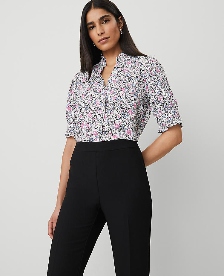A woman wearing a petite floral blouse with a ruffle collar, short puff sleeves with button cuffs, and a shirttail hem, featuring a floral pattern in pink, purple, and black on a white background.