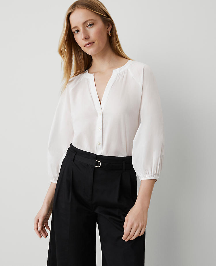 A woman wearing a white raglan popover top with blouson and raglan sleeves, split neck, tab collar, front buttons, and a hi-low shirttail hem.