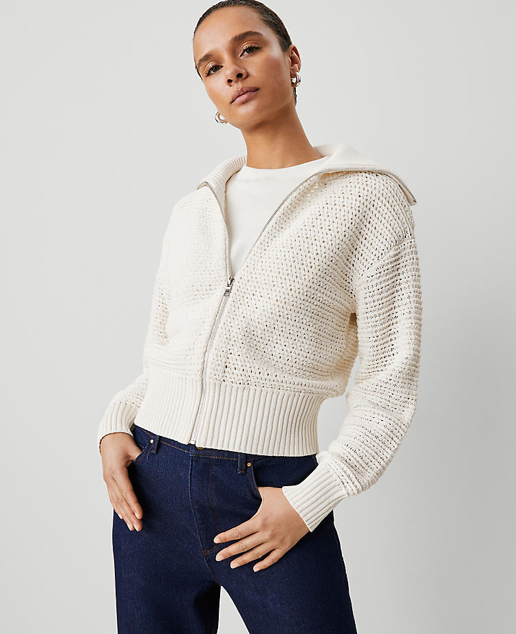 A woman wearing a cream open-stitch knit bomber jacket with a zip front, ribbed collar, cuffs, and hem, and long sleeves.