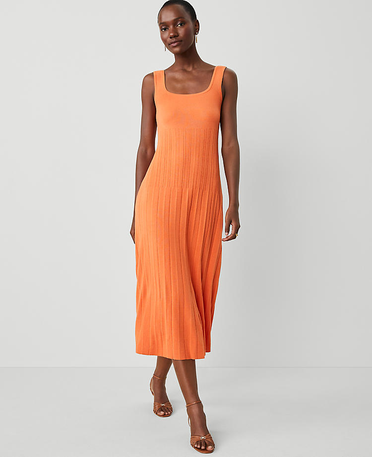 A woman wearing an orange pleated sleeveless sweater tank dress with a square neckline, standing against a plain background.