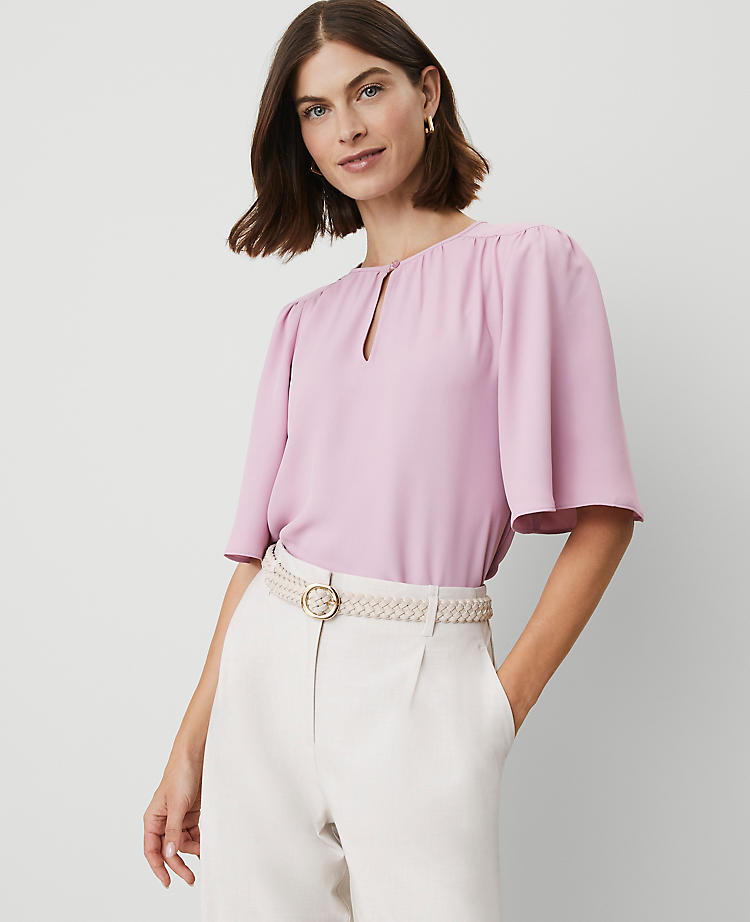A woman wearing a light pink petite draped sleeve blouse with a round neckline, short sleeves, and a shirttail hem.