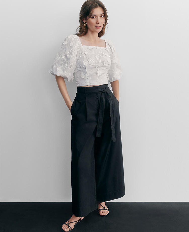 A woman wearing black cropped palazzo pants with a high waist, belt loops, and a tie belt, paired with a white embroidered top with puffed sleeves.