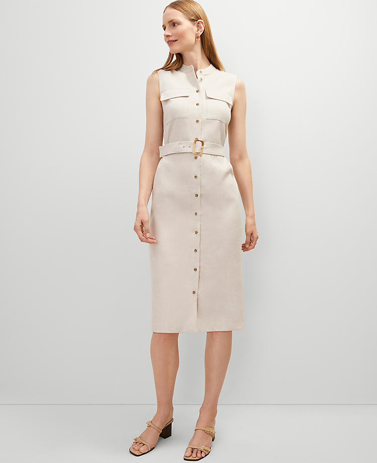 A woman wearing a sleeveless beige linen blend shirtdress with a stand collar, front buttons, flap pockets, and a removable belt.