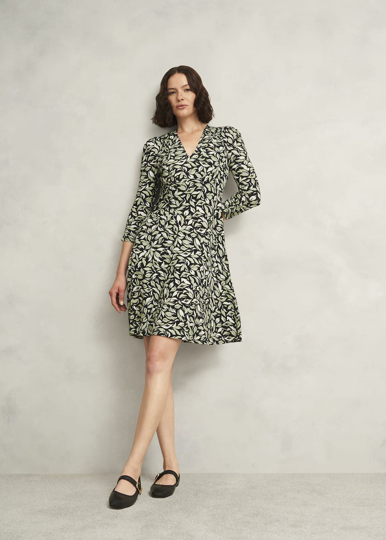 A woman wearing a petite Elosie Dress with a black and light green abstract leaf print, V-neckline, and long sleeves.