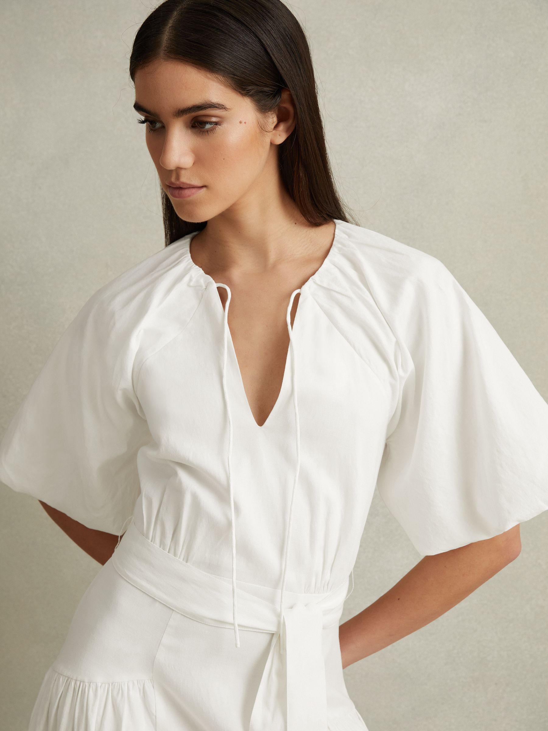 A woman wearing a white midi dress with puff sleeves, gathered waist, open-back, and a zip at the back.
