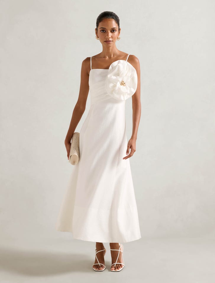 A woman wearing a white sleeveless midi dress with a square neckline, floral applique on the bodice, and a zip closure, paired with white high-heeled sandals and holding a clutch.
