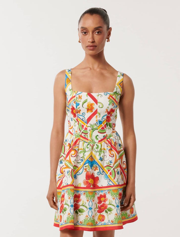 A woman wearing a colorful, patterned dress with a square neckline, wide straps, and a flared skirt, standing against a plain background.