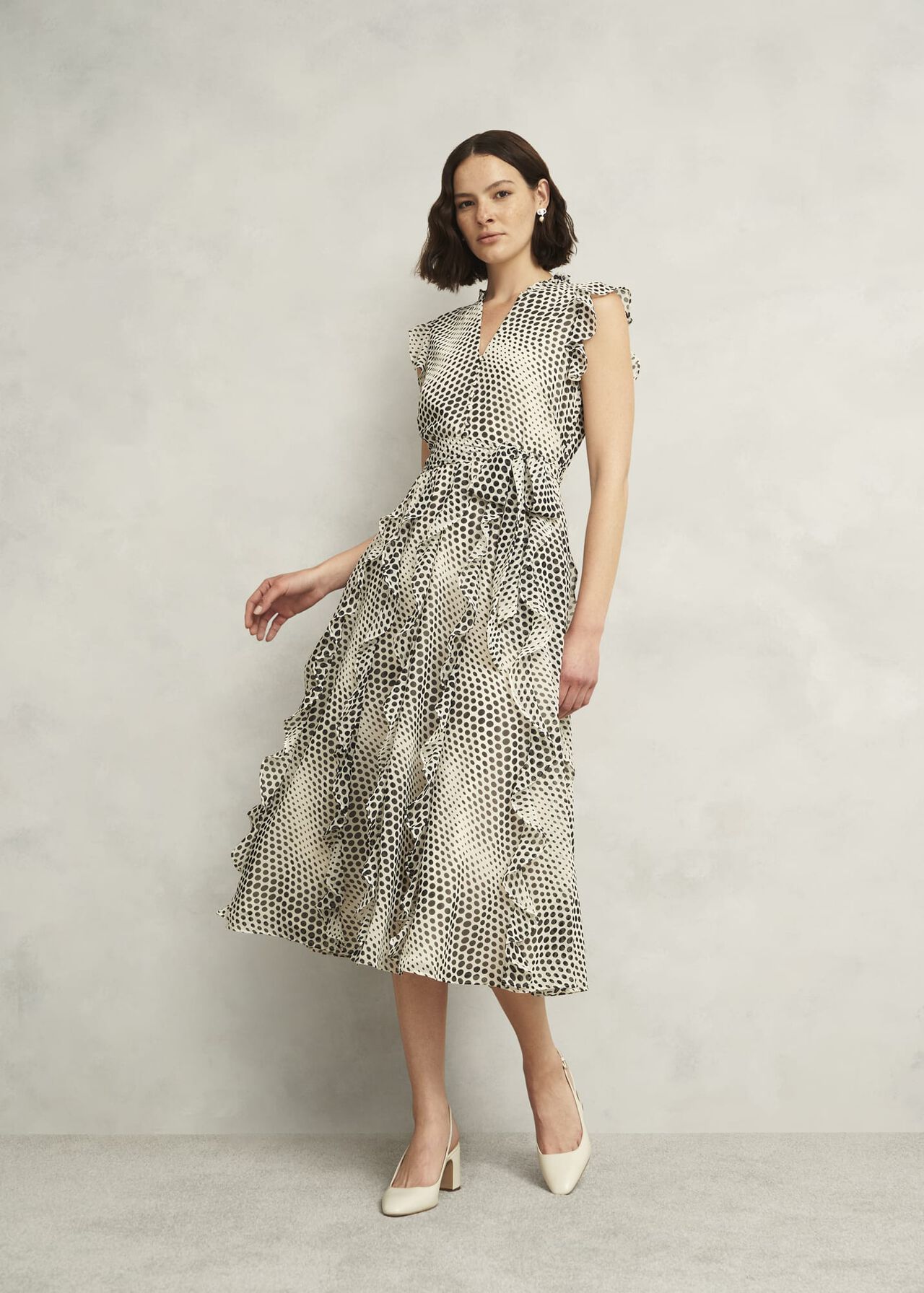 A woman wearing a beige midi dress with black polka dots, ruffle details on the sleeves and hem, a V-neckline, and a tie at the waist, standing against a plain background.