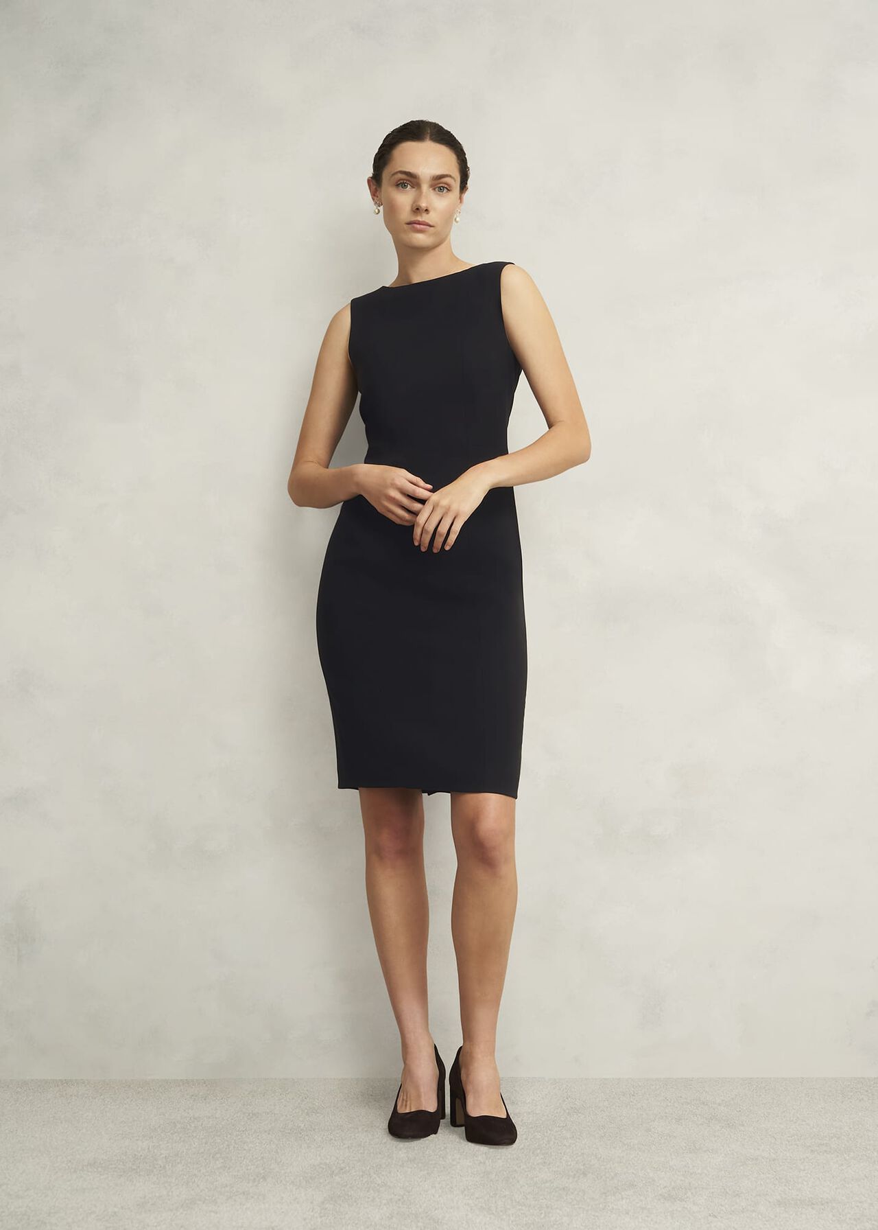 A woman wearing a black sleeveless shift dress with a boat neckline, standing against a plain background.