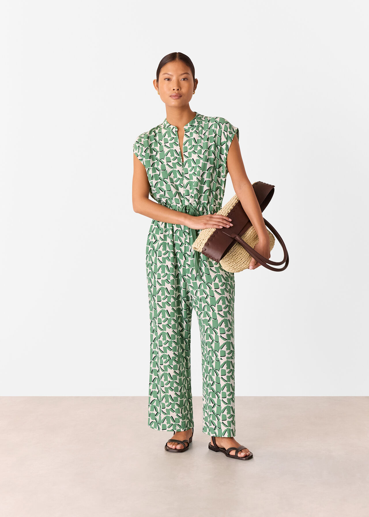 A woman wearing an ivory jumpsuit with a layered abstract squares pattern, short sleeves, and a relaxed-leg fit, holding a woven and leather handbag.