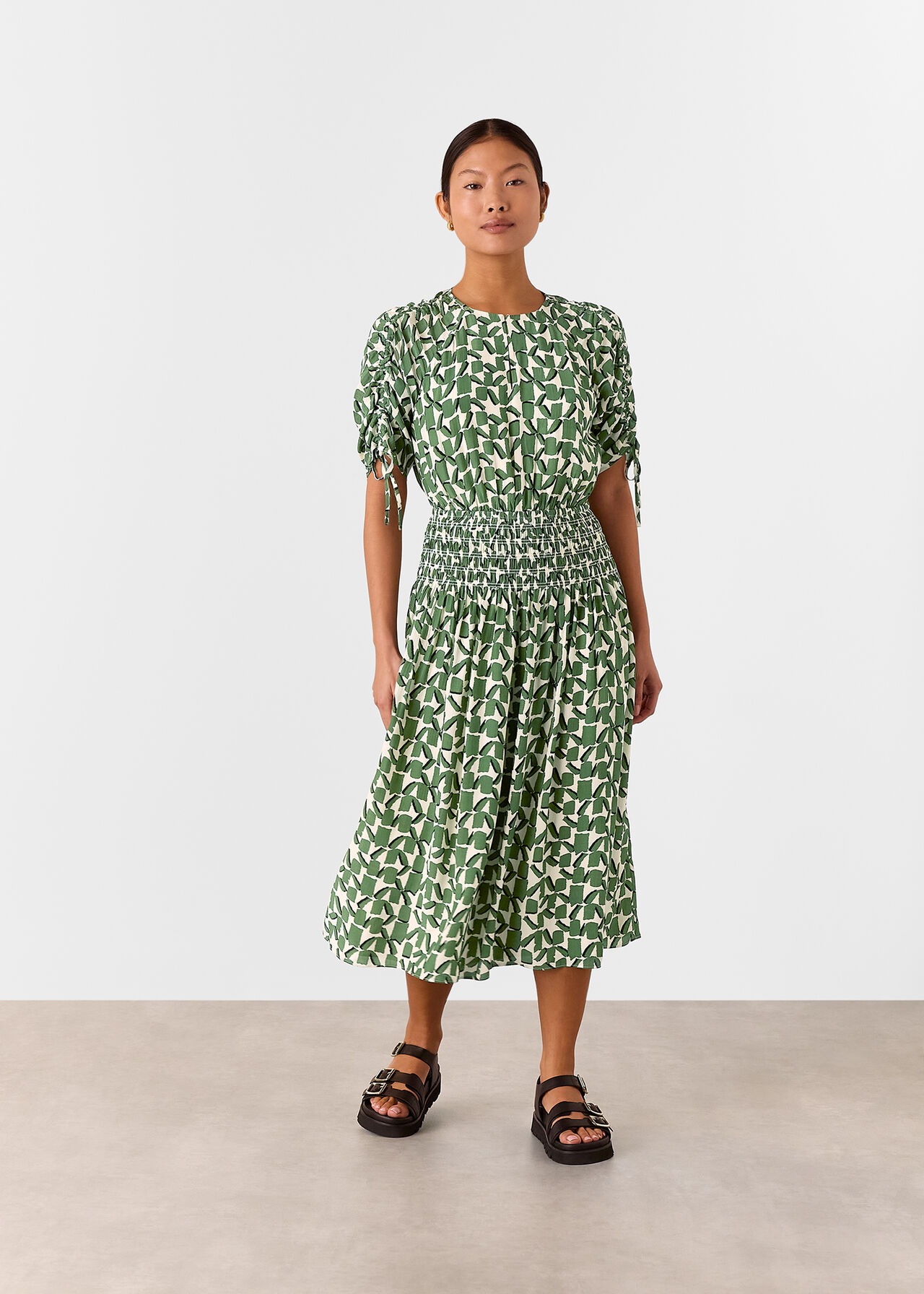 A woman wearing an ivory midi dress with green abstract square patterns, ruched short sleeves with drawstrings, a shirred waist, and a round neckline with a front keyhole.