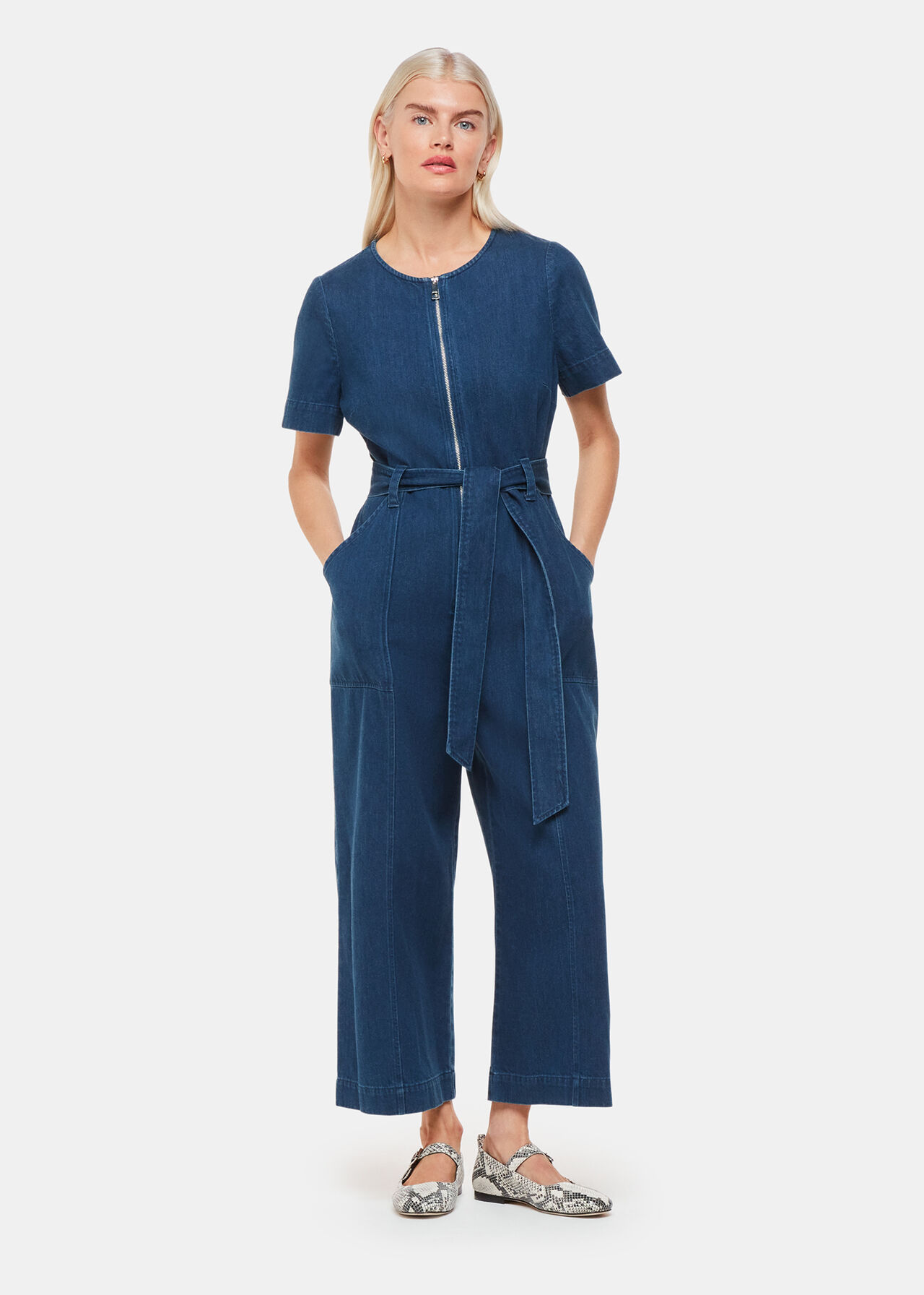 A woman wearing a blue petite short sleeve denim jumpsuit with a front zipper and a waist belt, standing with her hands in her pockets.