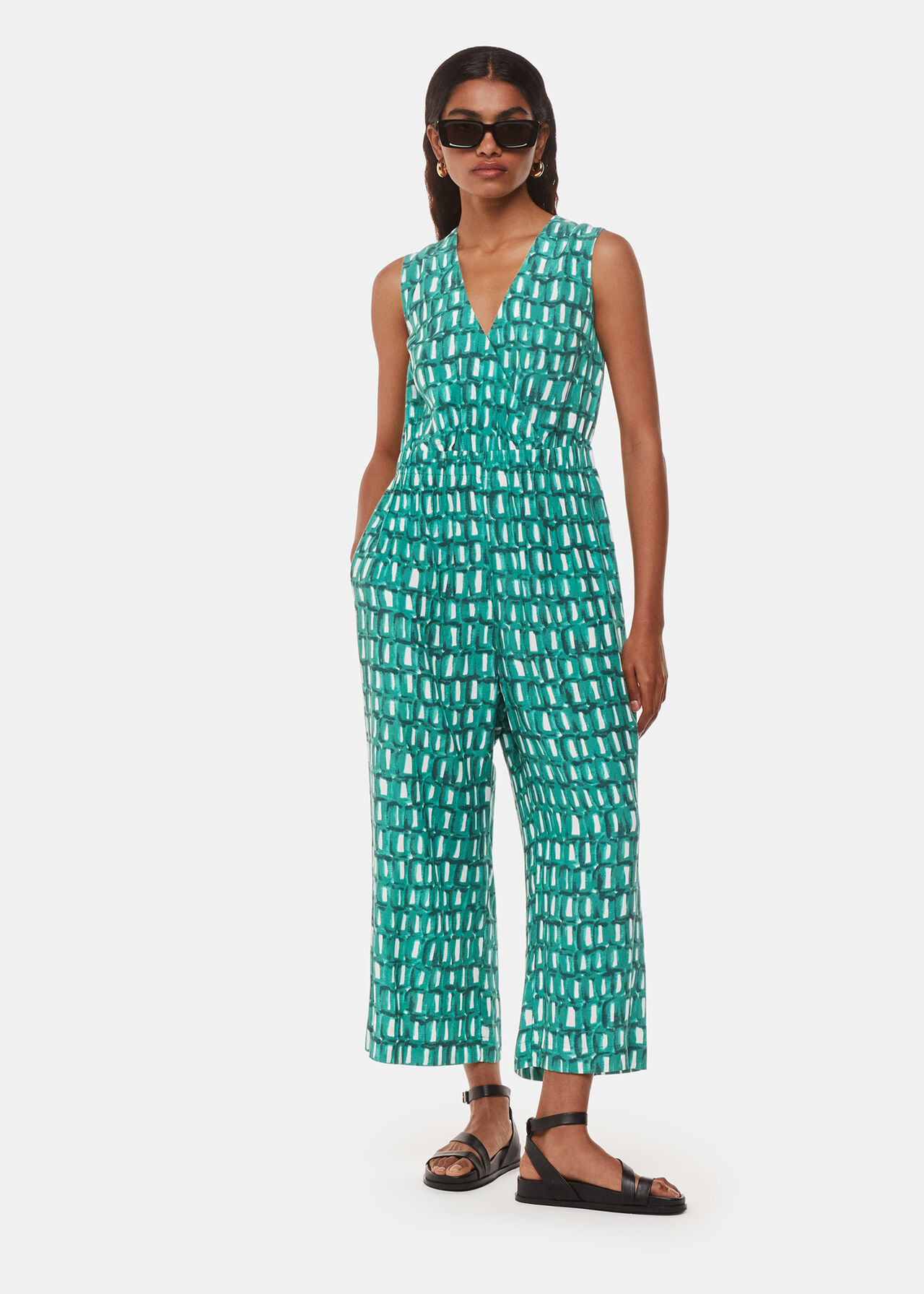 A woman wearing a green sleeveless jumpsuit with a V-neckline and a fitted waist, featuring a unique print, paired with black sandals and sunglasses.