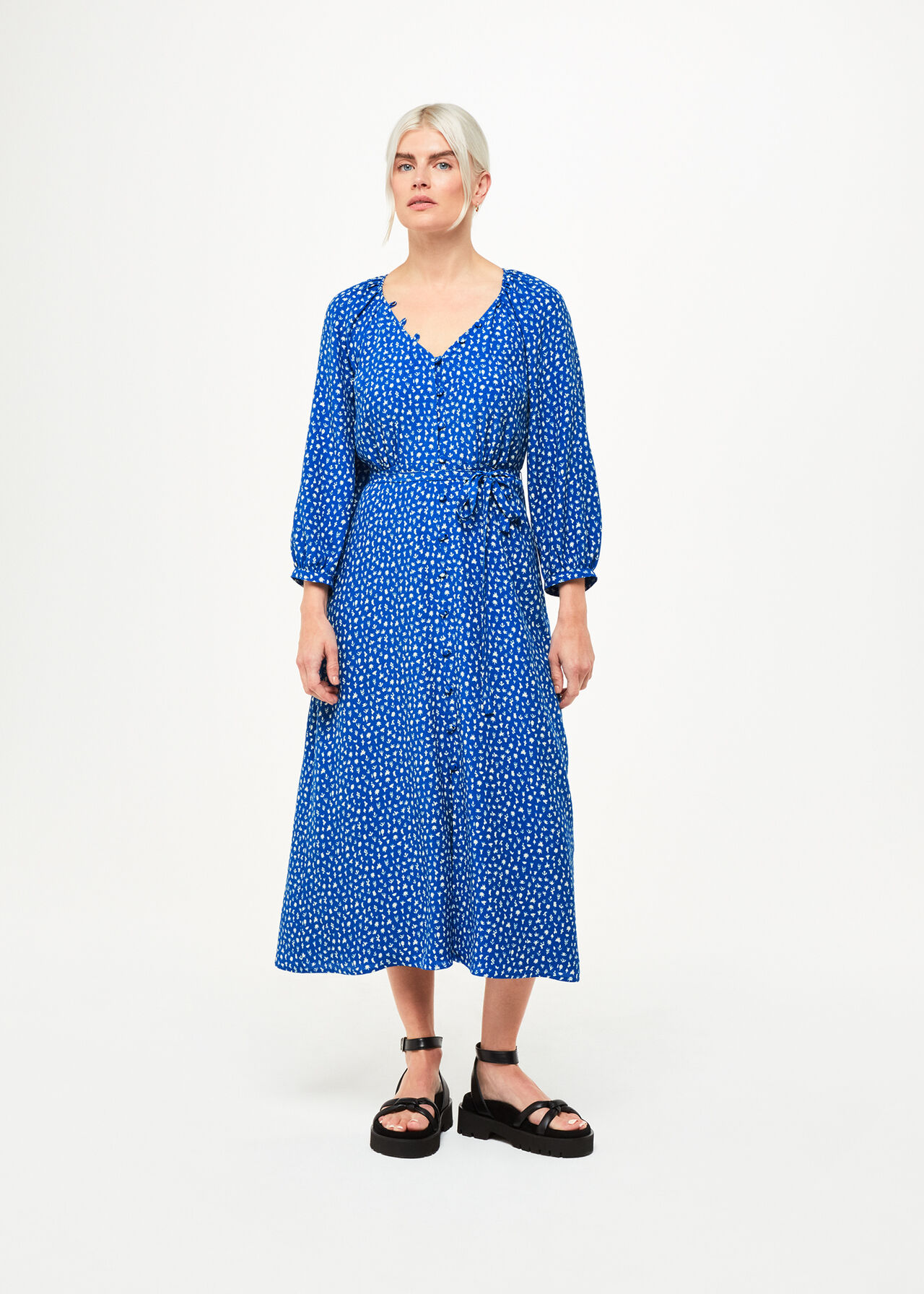 A woman wearing a blue midi dress with a micro cactus print, V-neckline, and waist tie, paired with black sandals.
