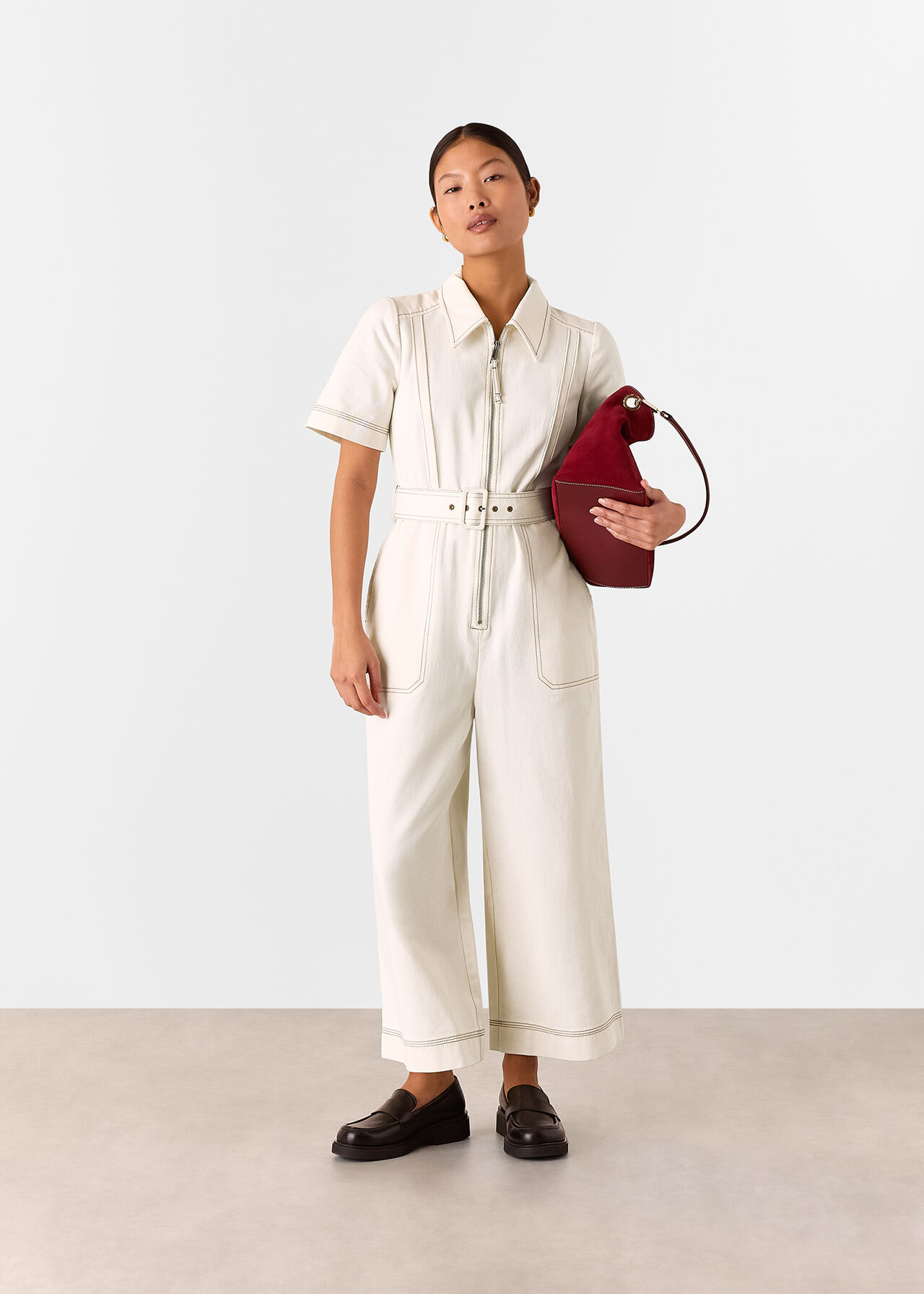 A woman wearing a fitted ivory denim jumpsuit with a belt, short sleeves, and a collared neckline, holding a red handbag.