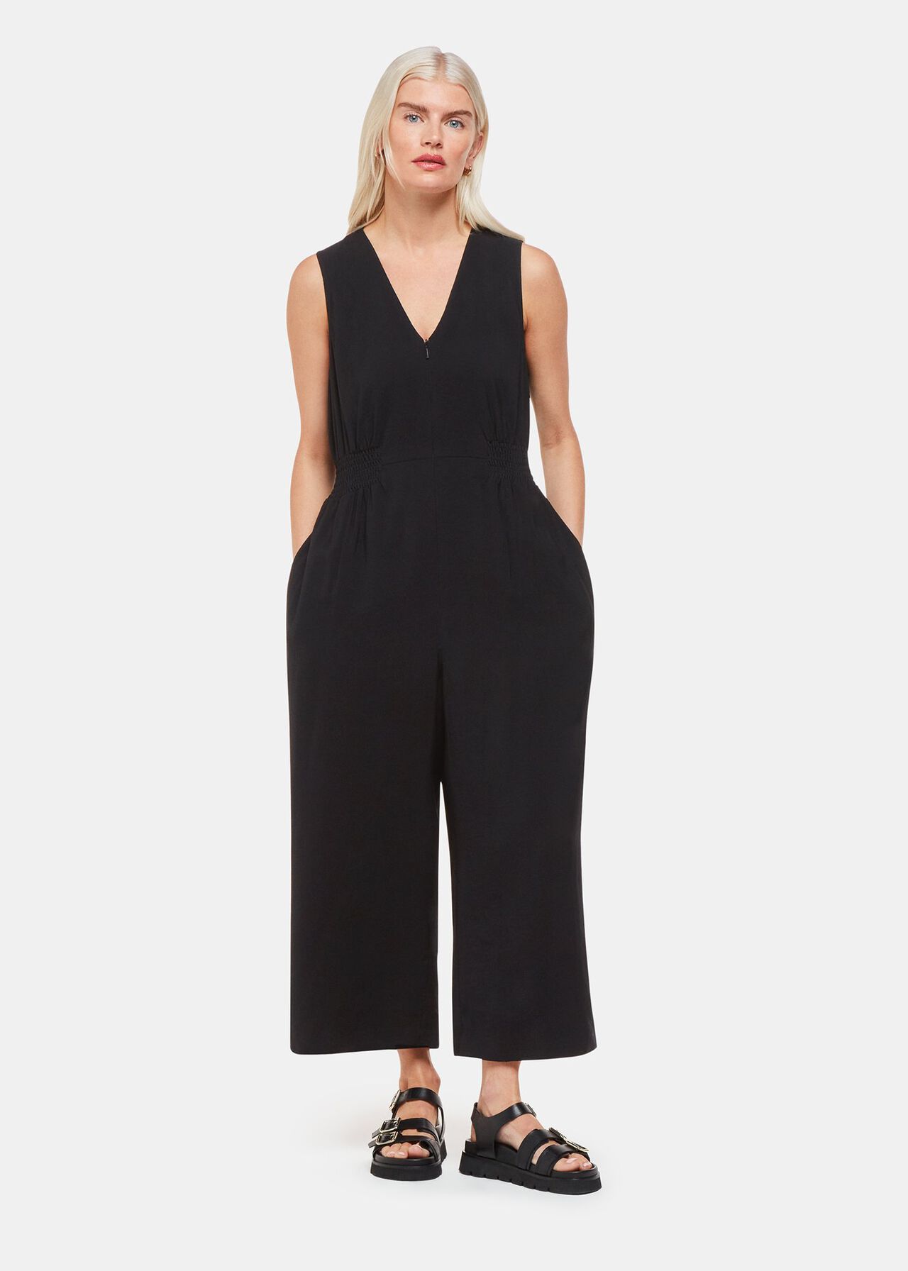 A woman wearing a black sleeveless jumpsuit with a V-neckline and cinched waist, standing against a plain background.
