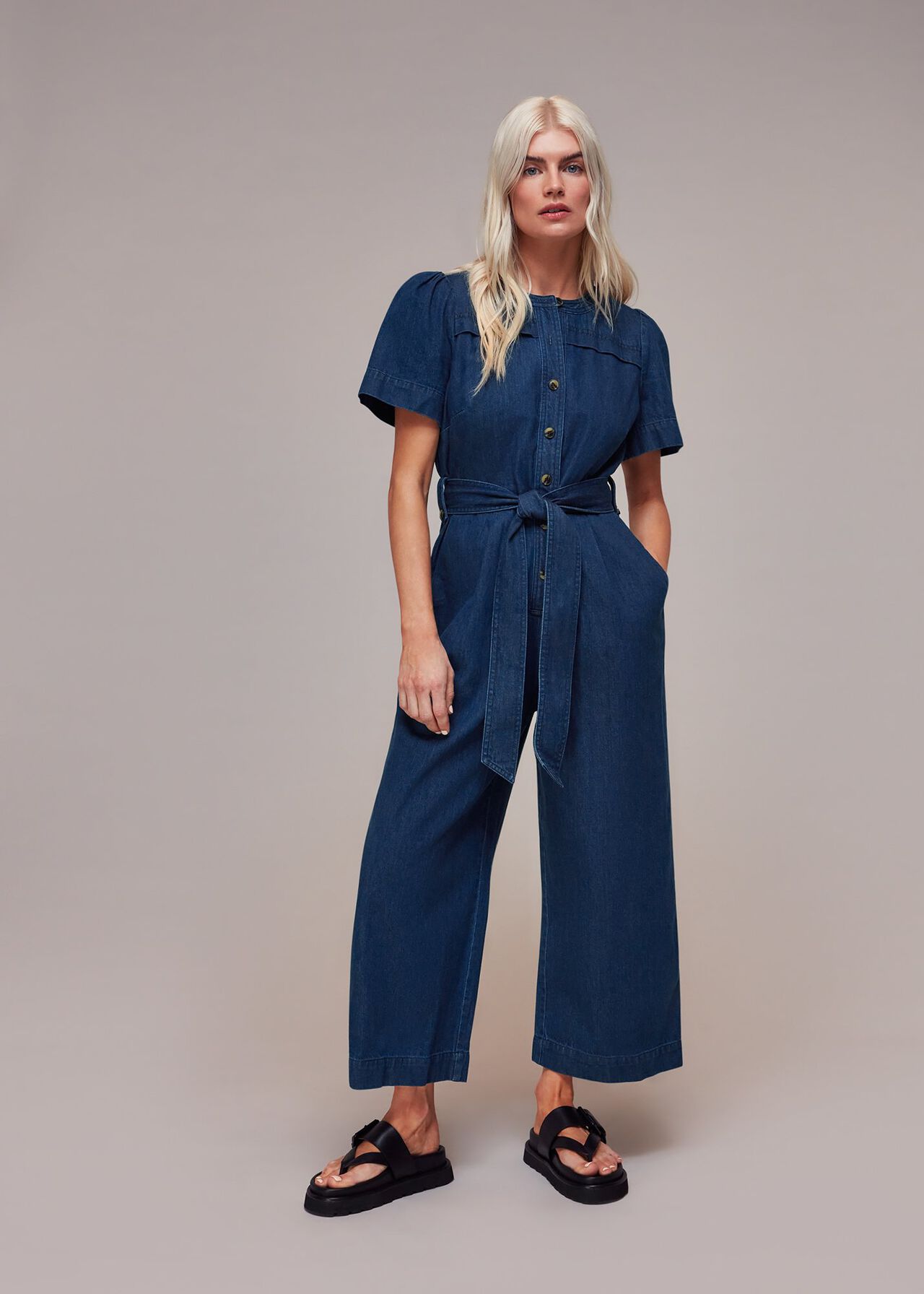 A woman wearing a blue denim jumpsuit with a tie waist belt, button-down front, and ankle-length hem, standing against a plain background.