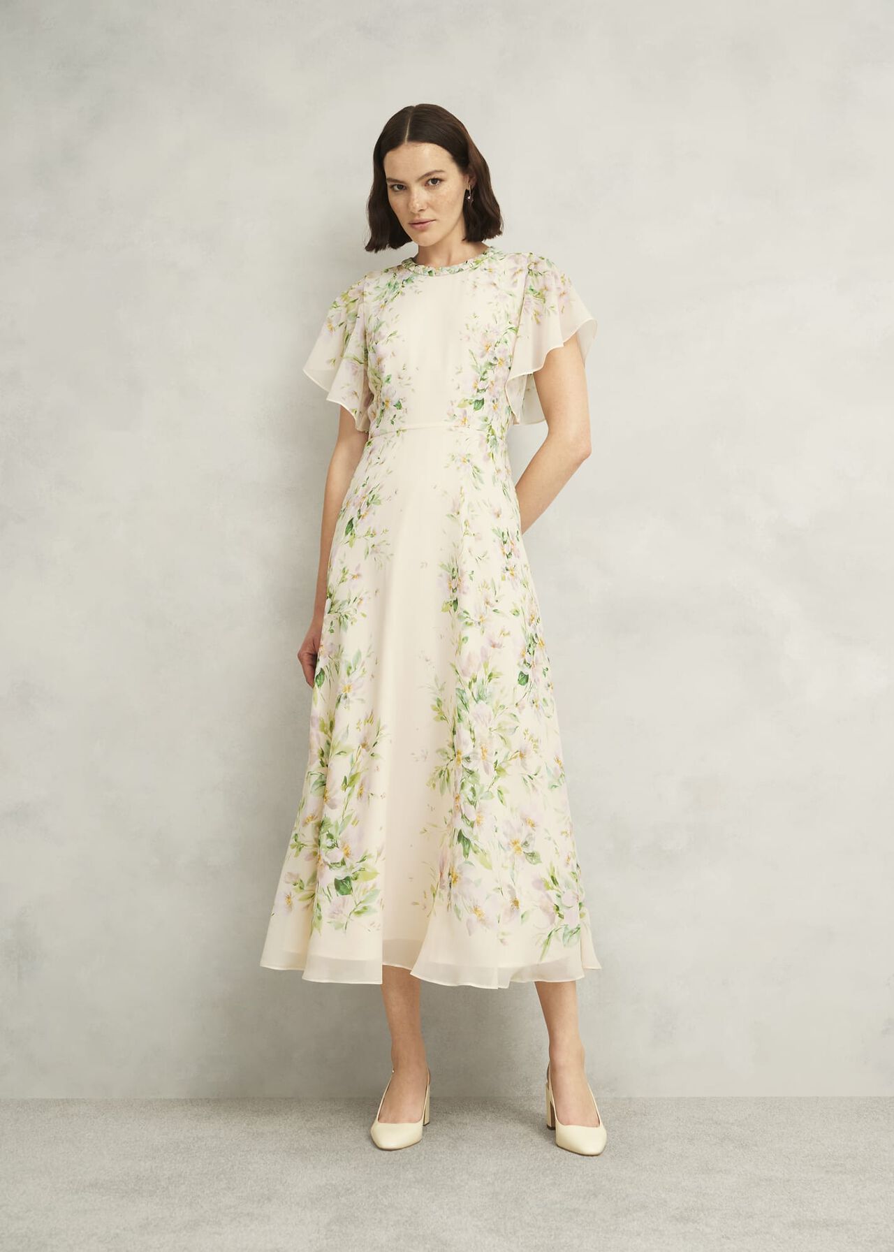 A woman wearing a cream-colored silk dress with a floral print, plaited neckline, waterfall sleeves, and a feminine silhouette.