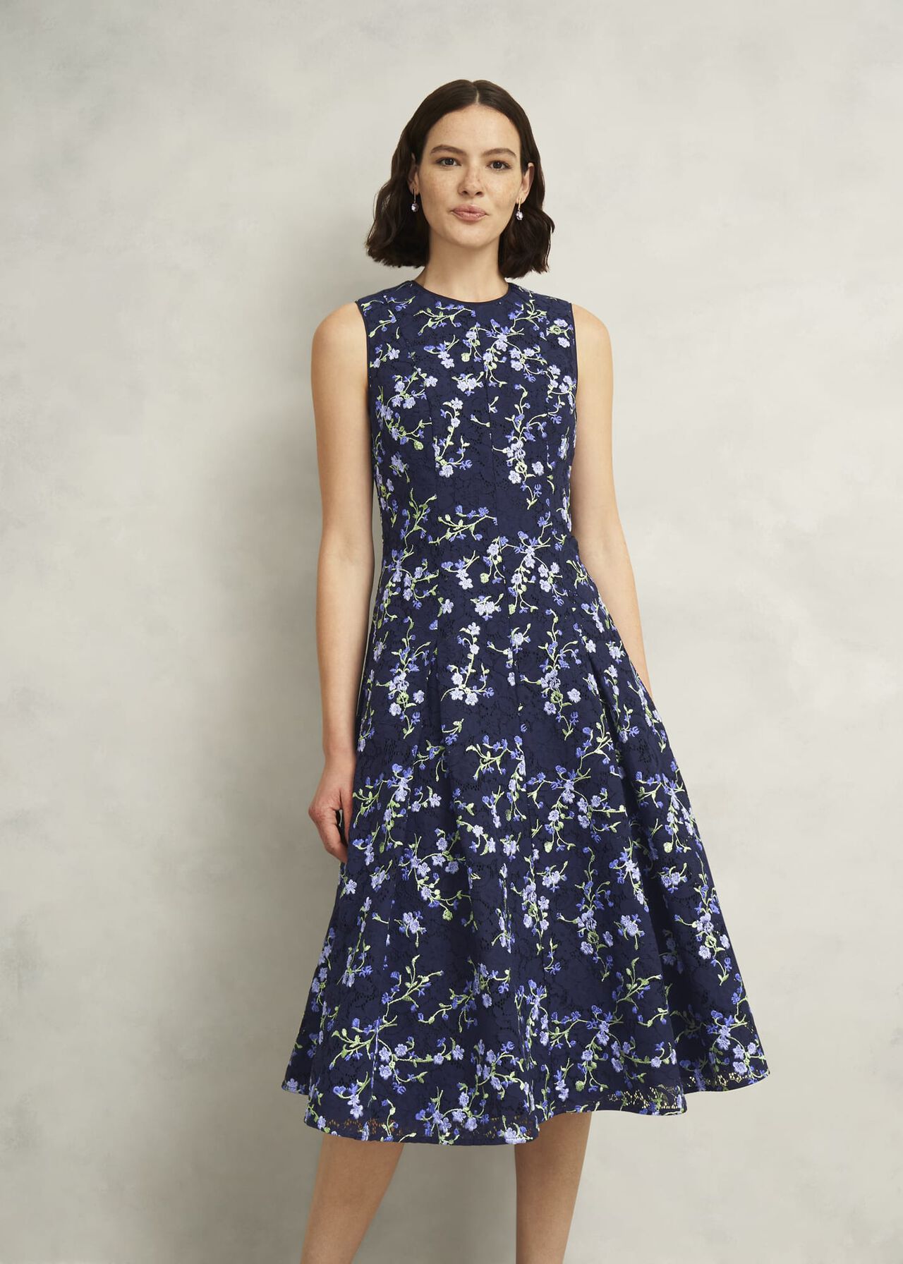 A woman wearing a navy blue sleeveless embroidered floral dress with lace details, pleated skirt, and side pockets.