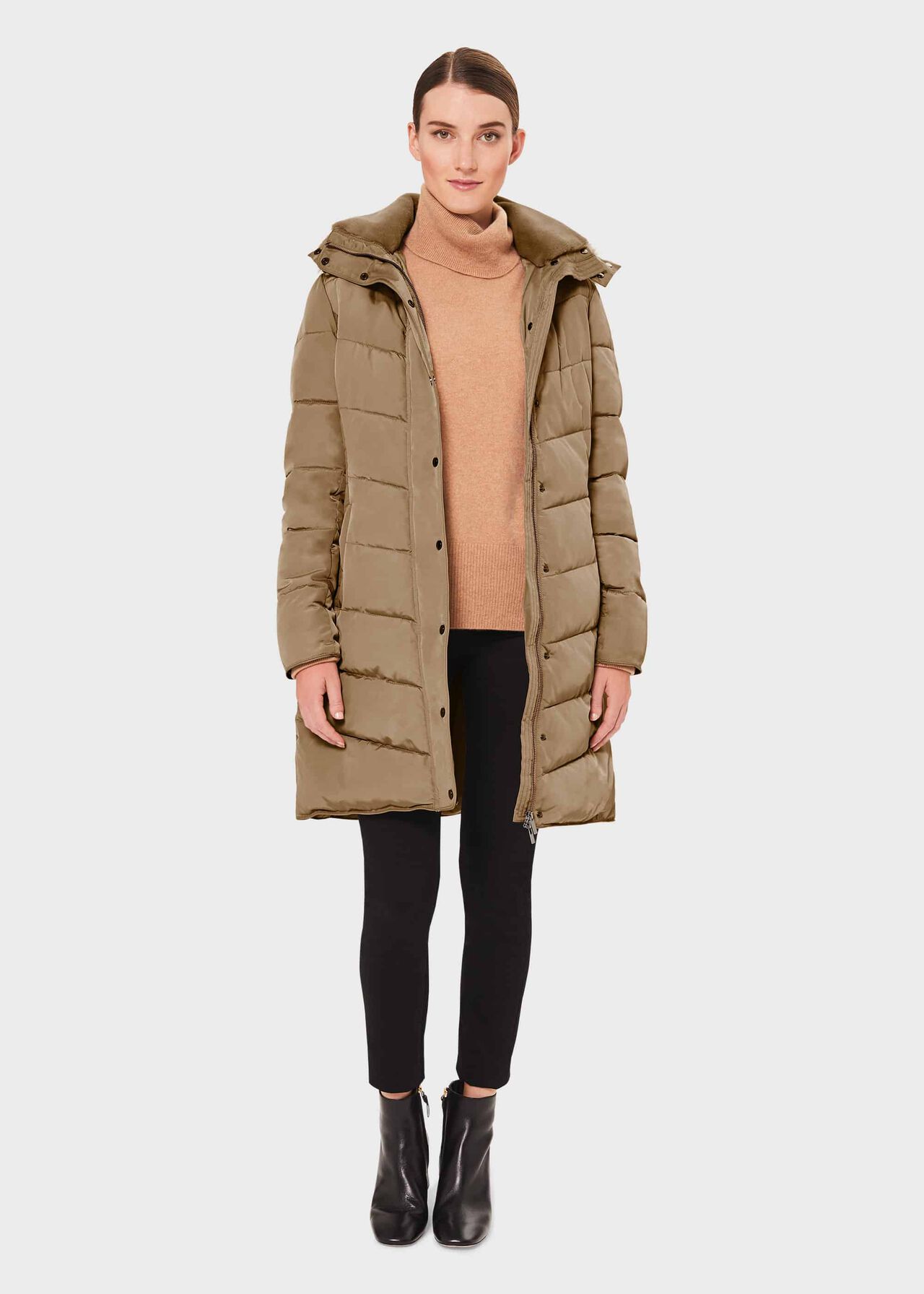 A woman wearing a beige puffer jacket with a hood, standing against a plain background.