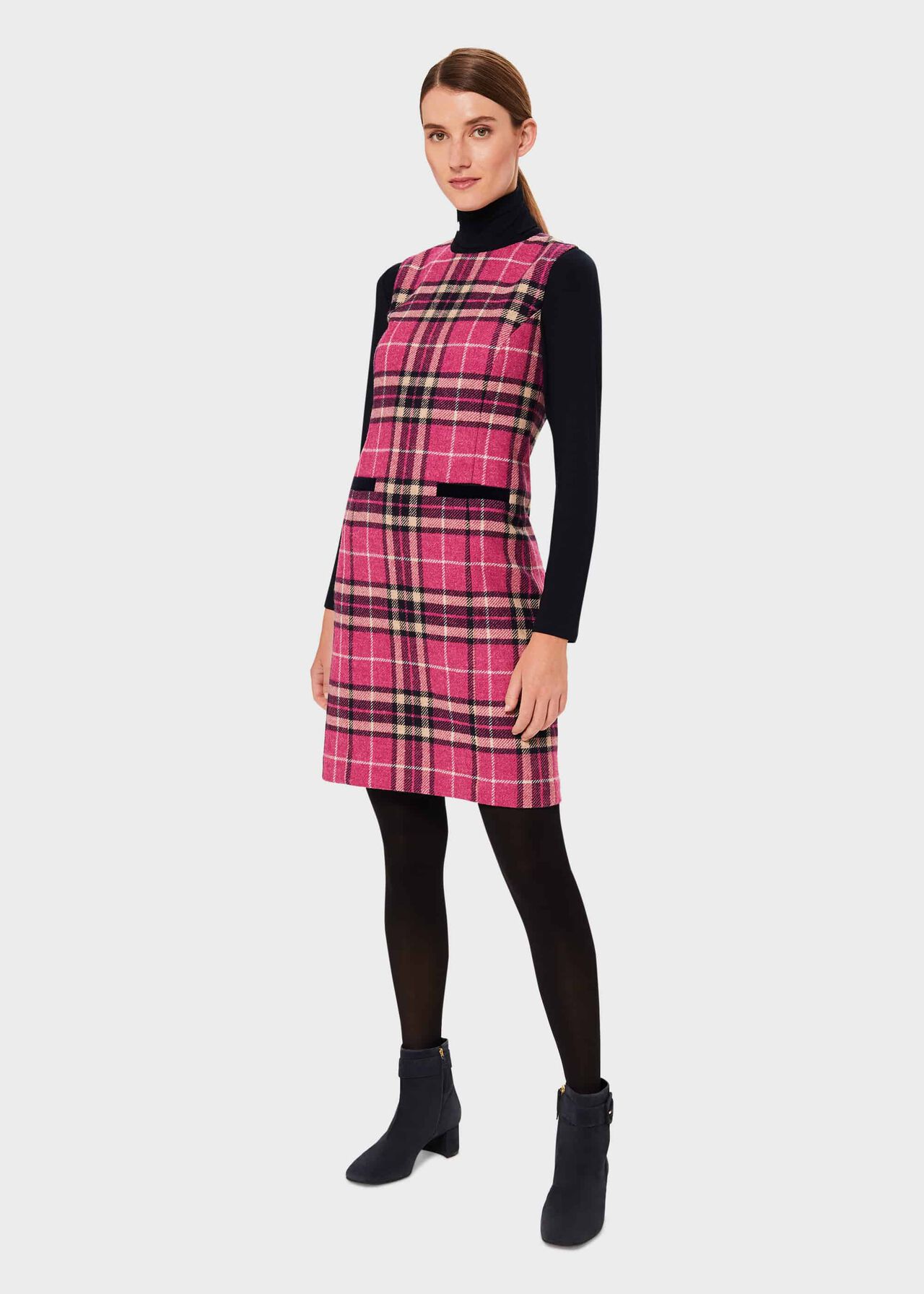 A woman wearing a pink check pattern wool shift dress with black long sleeves and black tights, paired with black ankle boots.
