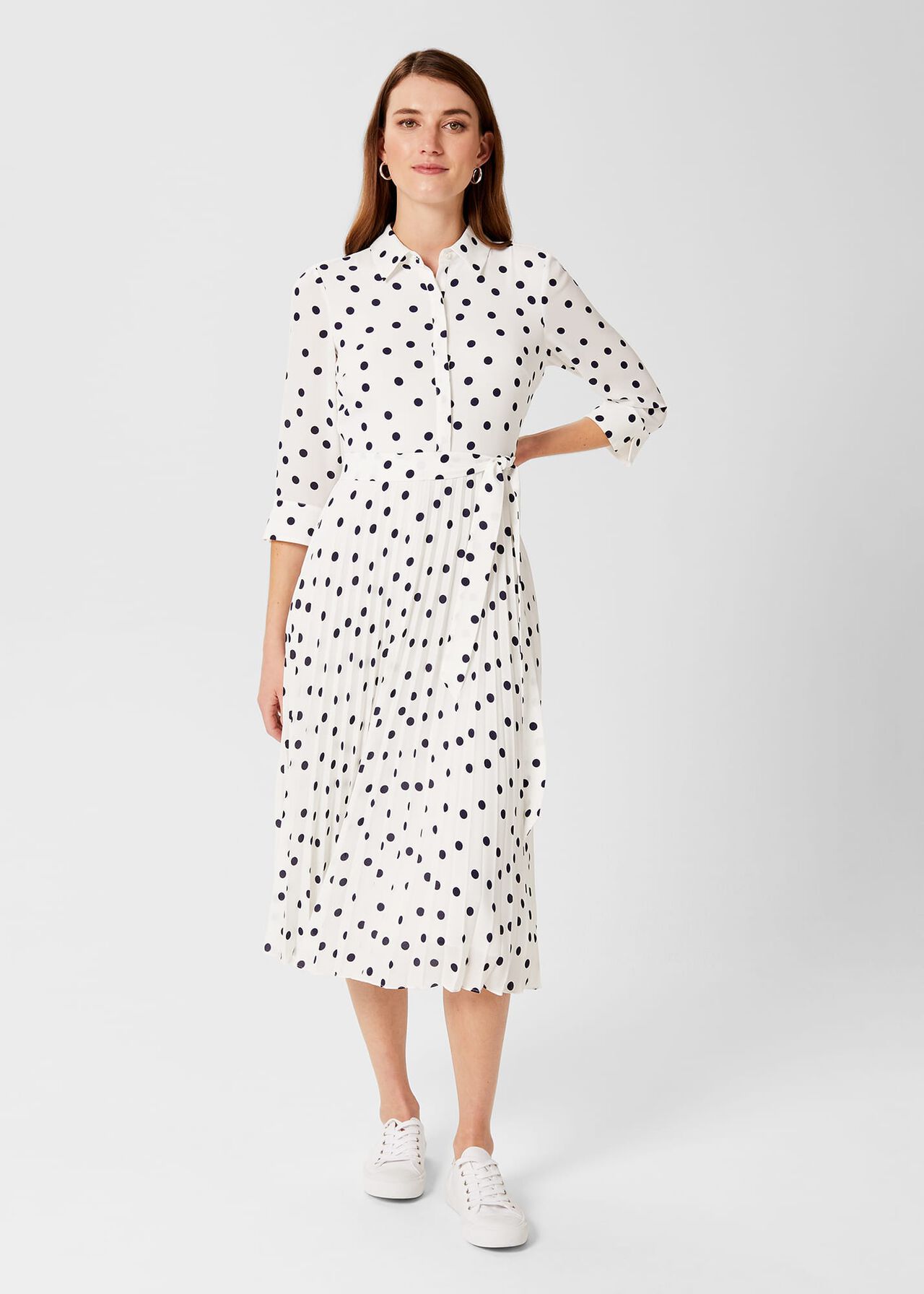 A woman wearing a white midi dress with black polka dots, pleated skirt, tie belt, and collared neckline, standing against a plain background.