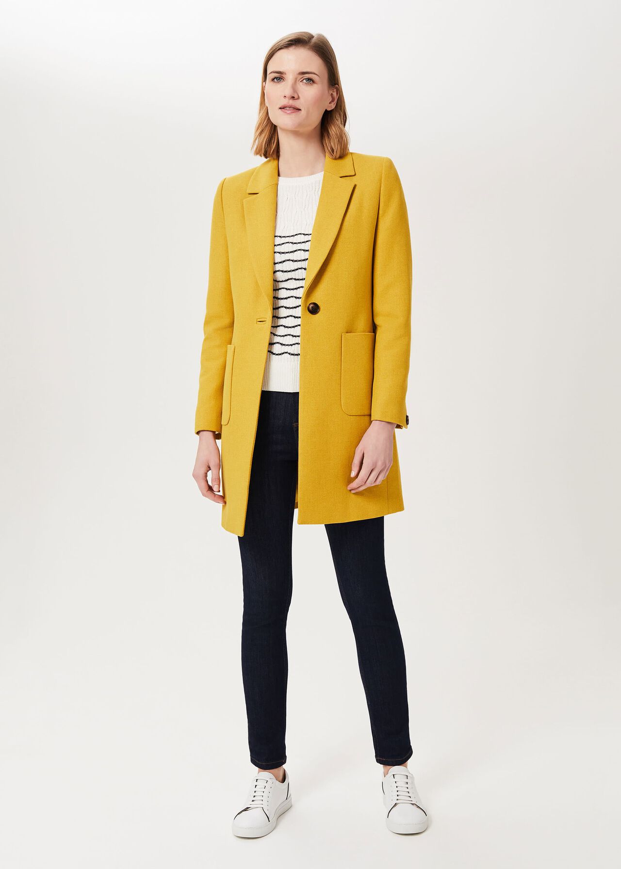A woman wearing a yellow petite Corina coat with a collar, patch pockets, and a contrast felt undercollar, styled with a white and black striped sweater, dark jeans, and white sneakers.