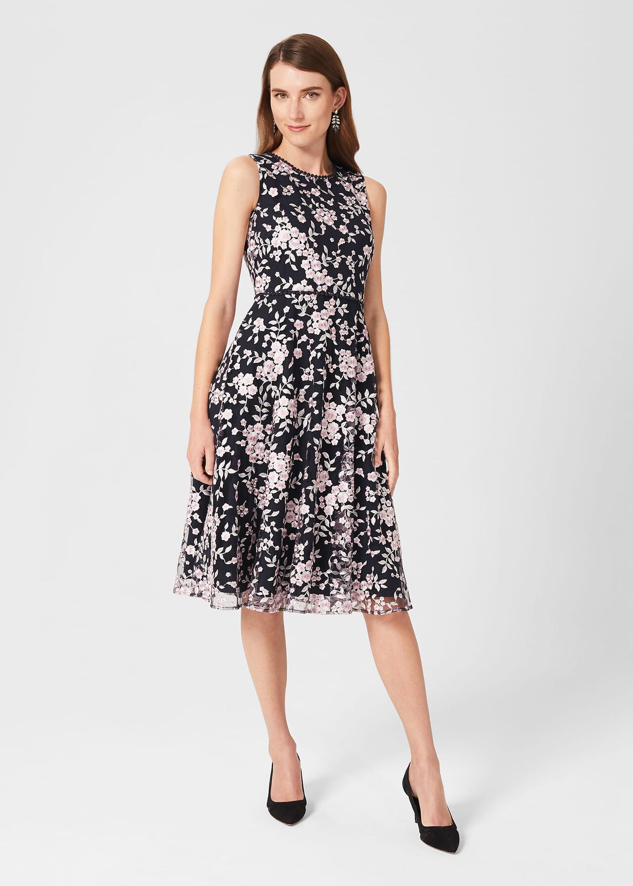 A woman wearing a black floral embroidered dress with a fit and flare silhouette, circle trim on the neck and waist, and a midi length.