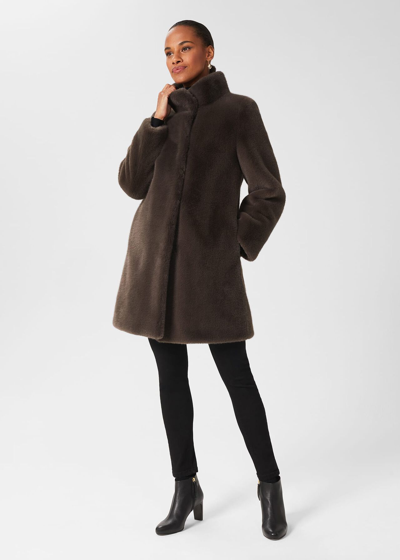 A woman wearing a dark brown plush faux fur coat with a funnel neck, tailored fit, and mid-thigh length, styled with black pants and ankle boots.