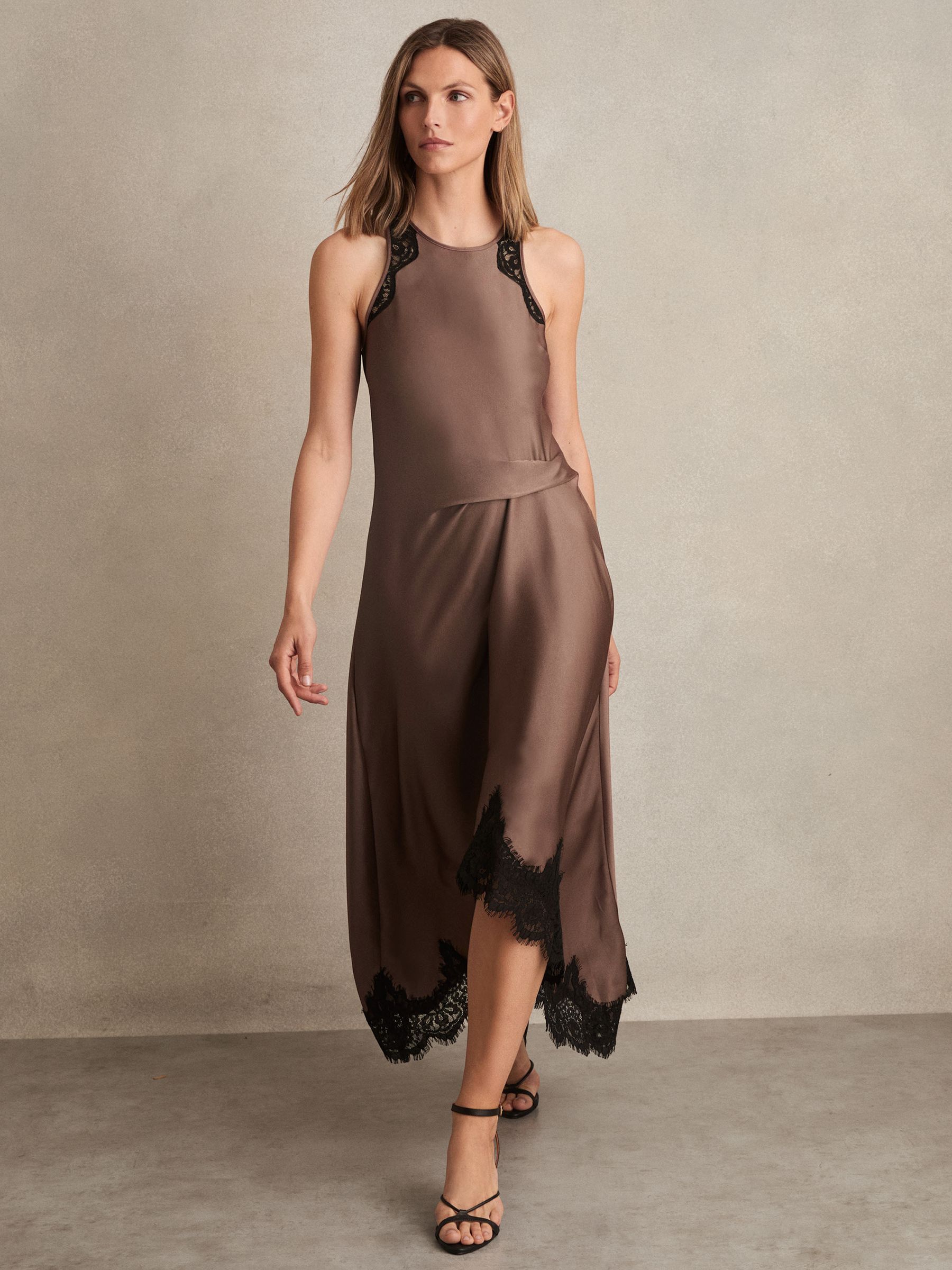 A woman wearing a mocha brown satin dress with lace trims at the armholes and hem, featuring a draped, asymmetric midi skirt, and a back button keyhole.
