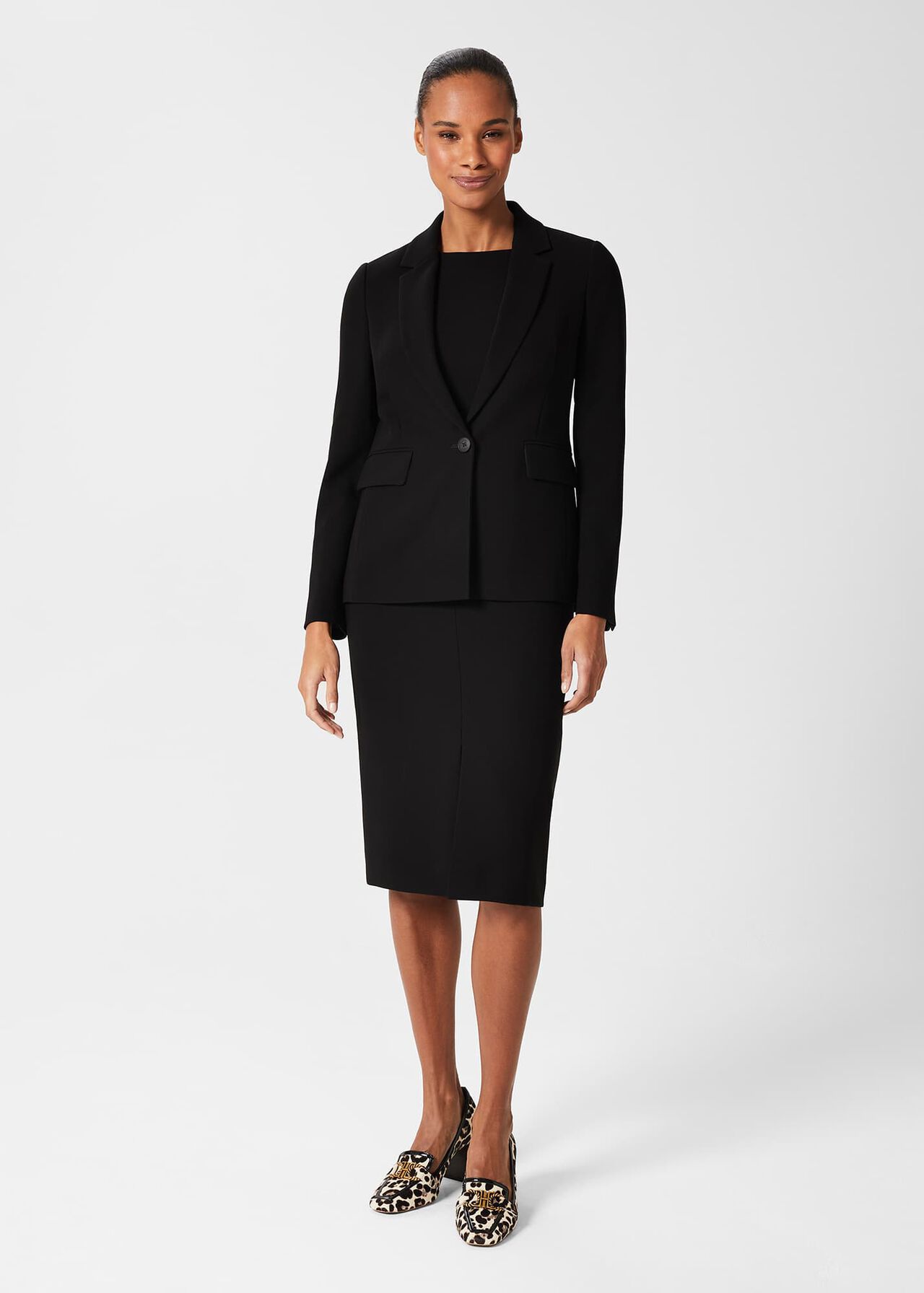 A woman wearing a black, longline women's jacket with notch lapels, flap pockets, and a collar, paired with a matching skirt and leopard print shoes.