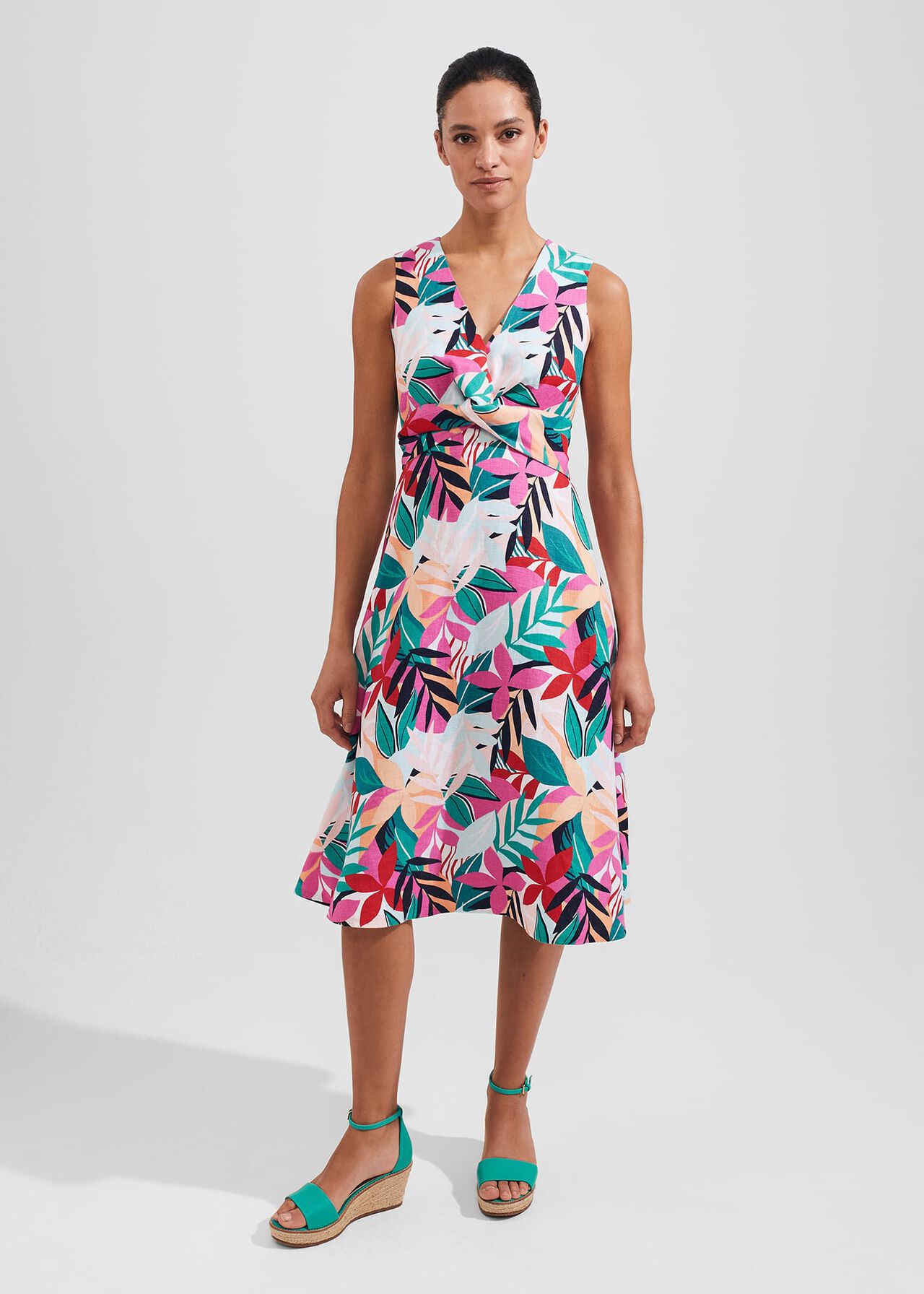 A woman wearing a sleeveless, colorful palm leaf print dress with a V-neckline, twist waist, and side pockets.