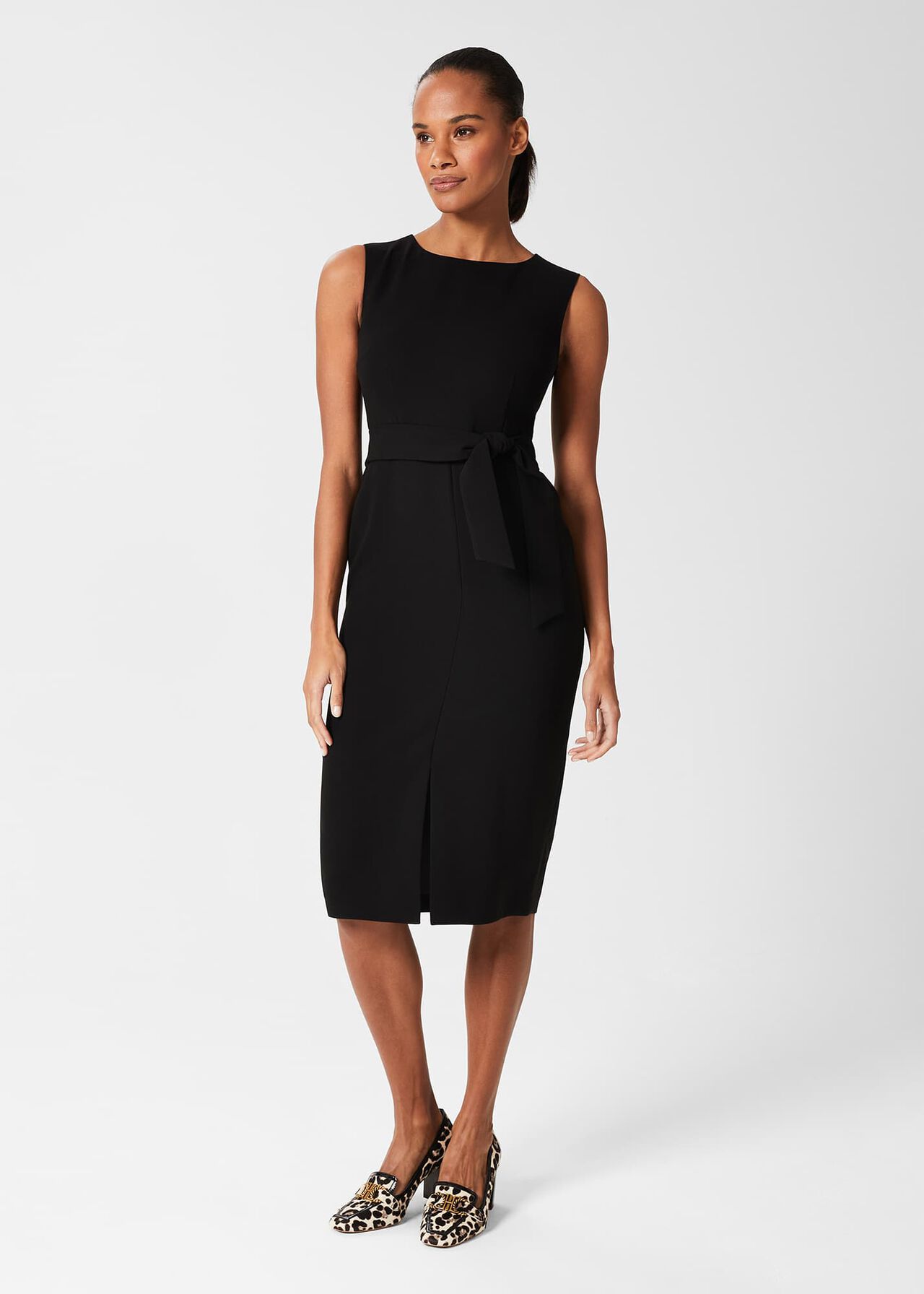 A woman wearing a sleeveless black belted dress with a crew neck and front split, paired with leopard print heels.