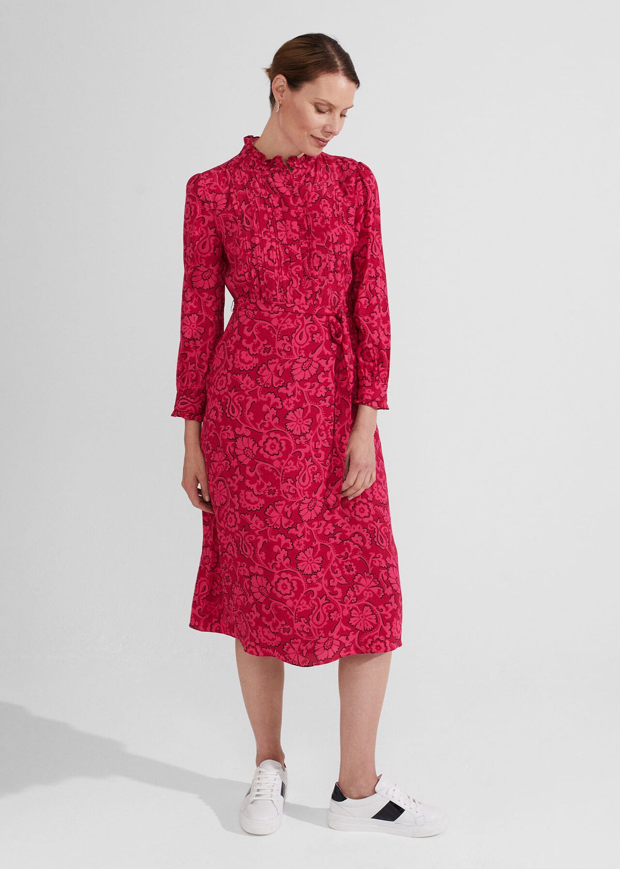 A woman wearing a pink floral print dress with a frill collar, long sleeves with elasticated cuffs, and a tie belt, paired with white sneakers.