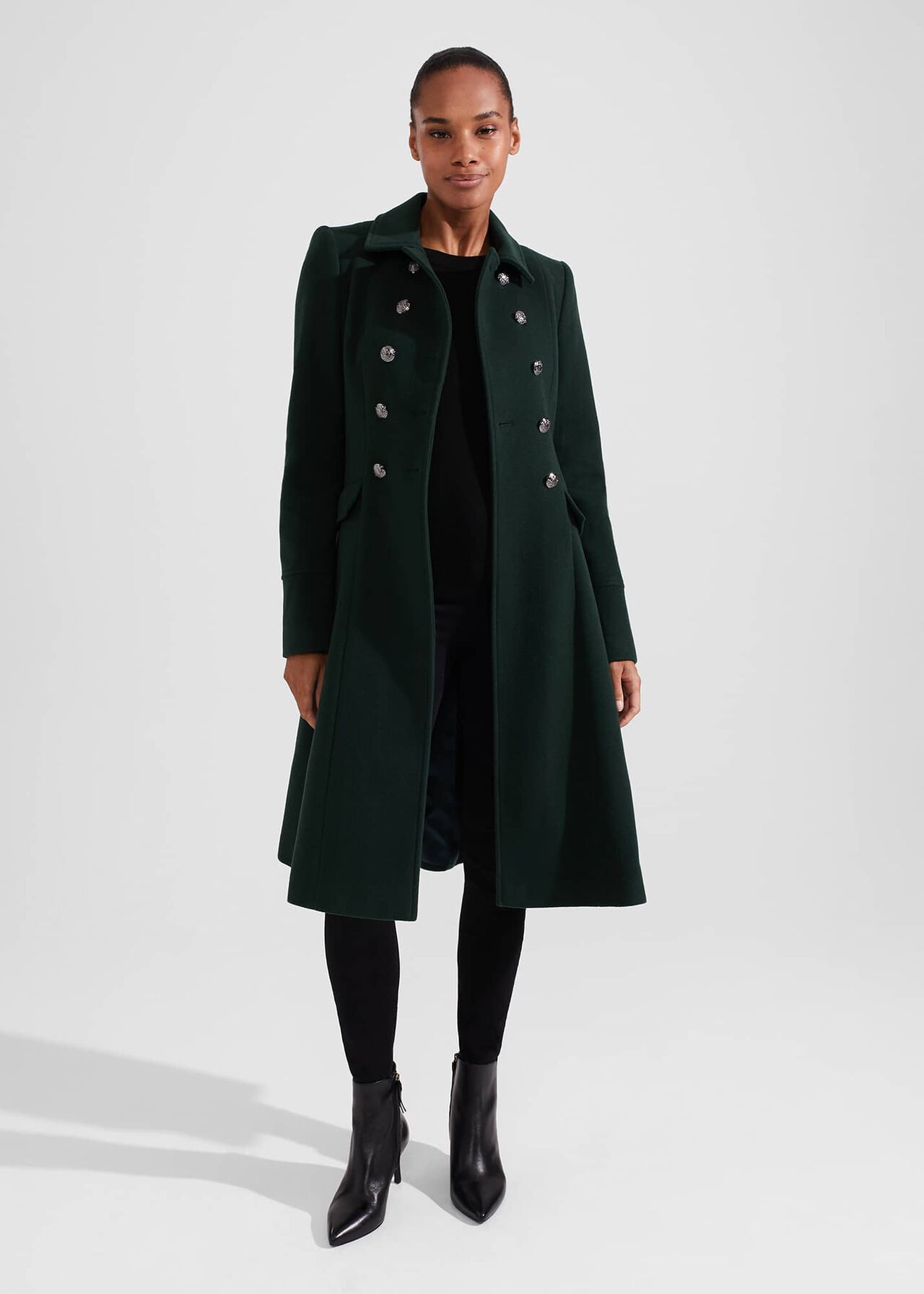 A woman wearing a dark green wool blend coat with a collar, front buttons, and ticket pockets, styled with black pants and boots.