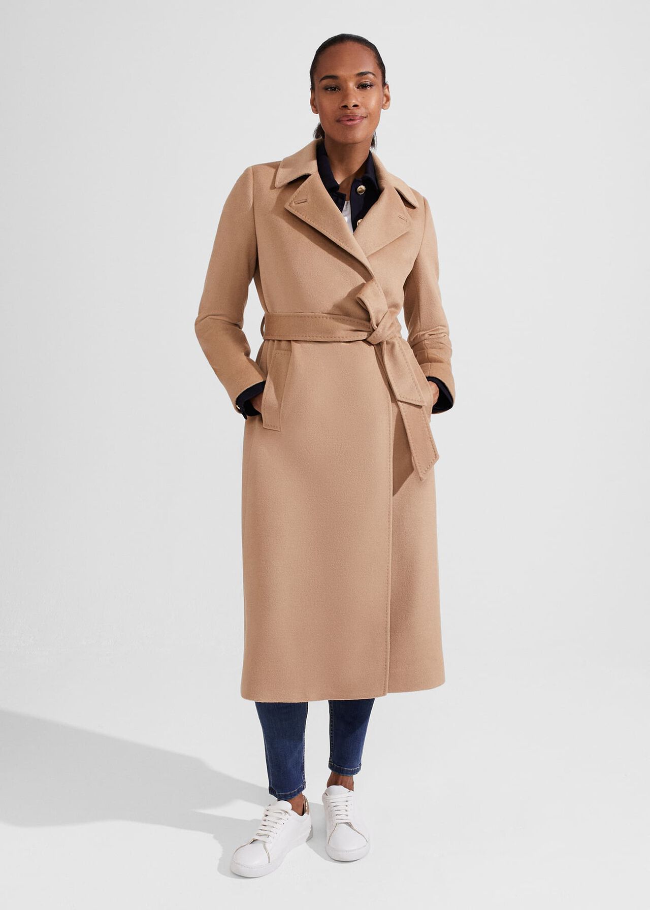 A woman wearing a beige, tailored, midi-length coat with a belt, slip pockets, and a button under the collar.