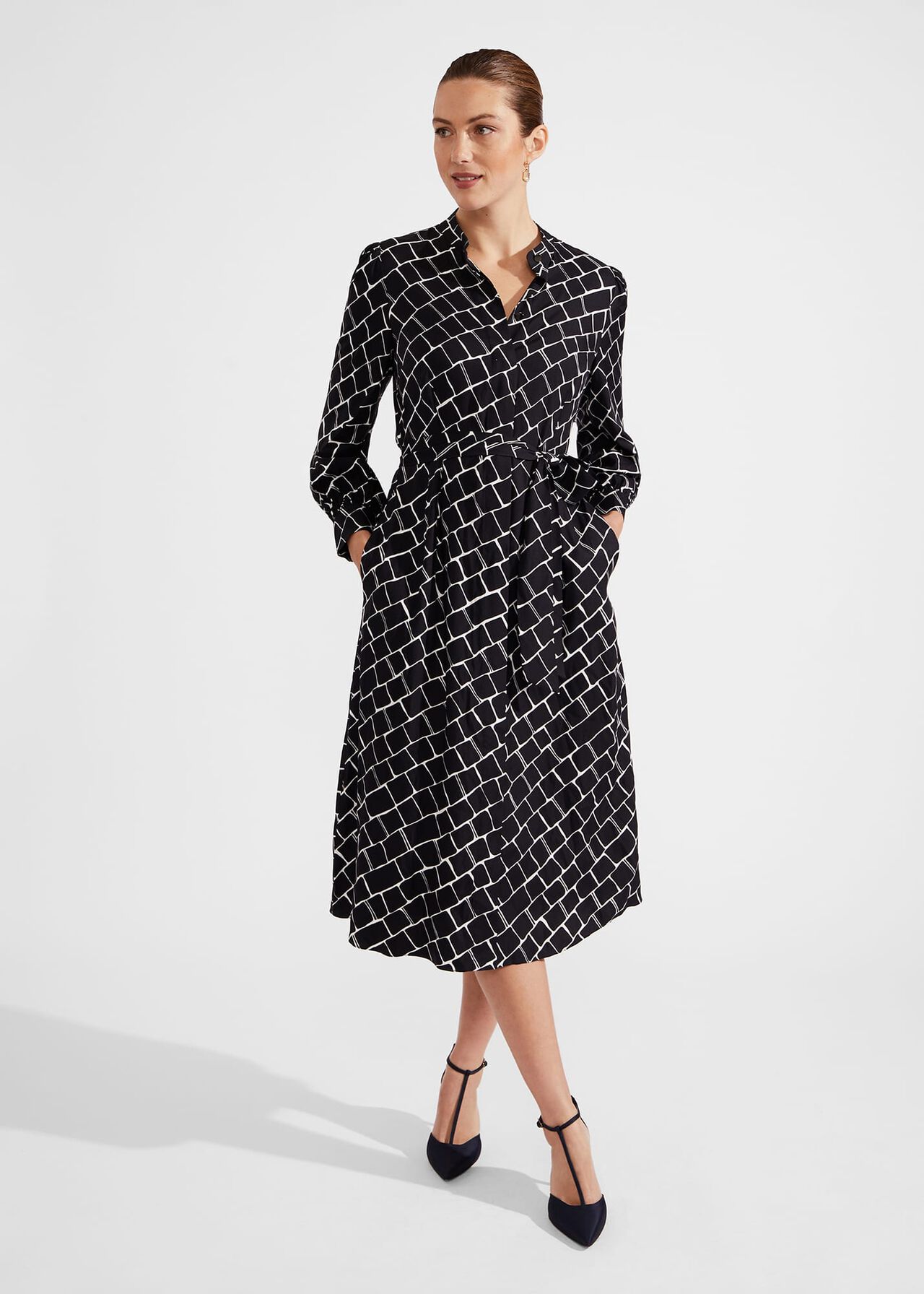 A woman wearing a black and white tile print midi dress with a tie waist belt, side pockets, mandarin collar, and covered placket.