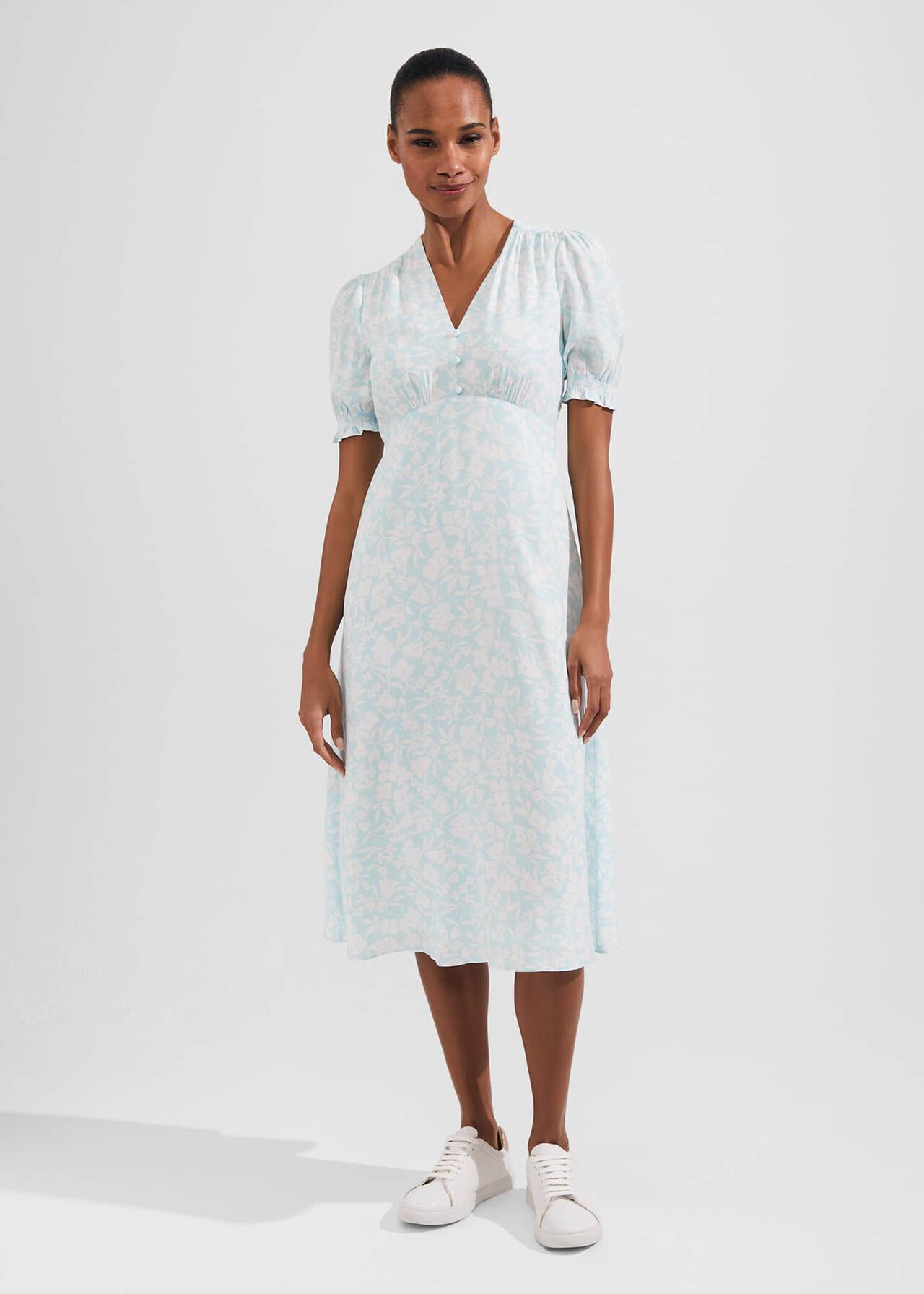 A woman wearing a light blue midi dress with floral pattern, short puff sleeves with frill cuffs, V-neckline, and front buttons.