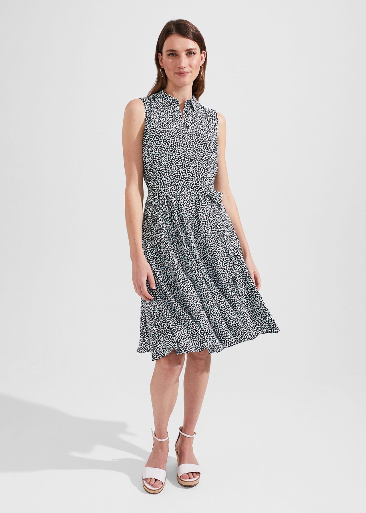 A woman wearing a sleeveless black and white spot print dress with a collar, tie belt, and side pockets.