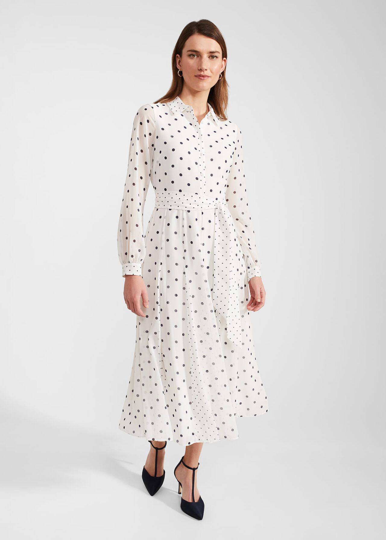 A woman wearing a white midi dress with black polka dots, collar neckline, long sleeves with cuffs, and a tie waist.