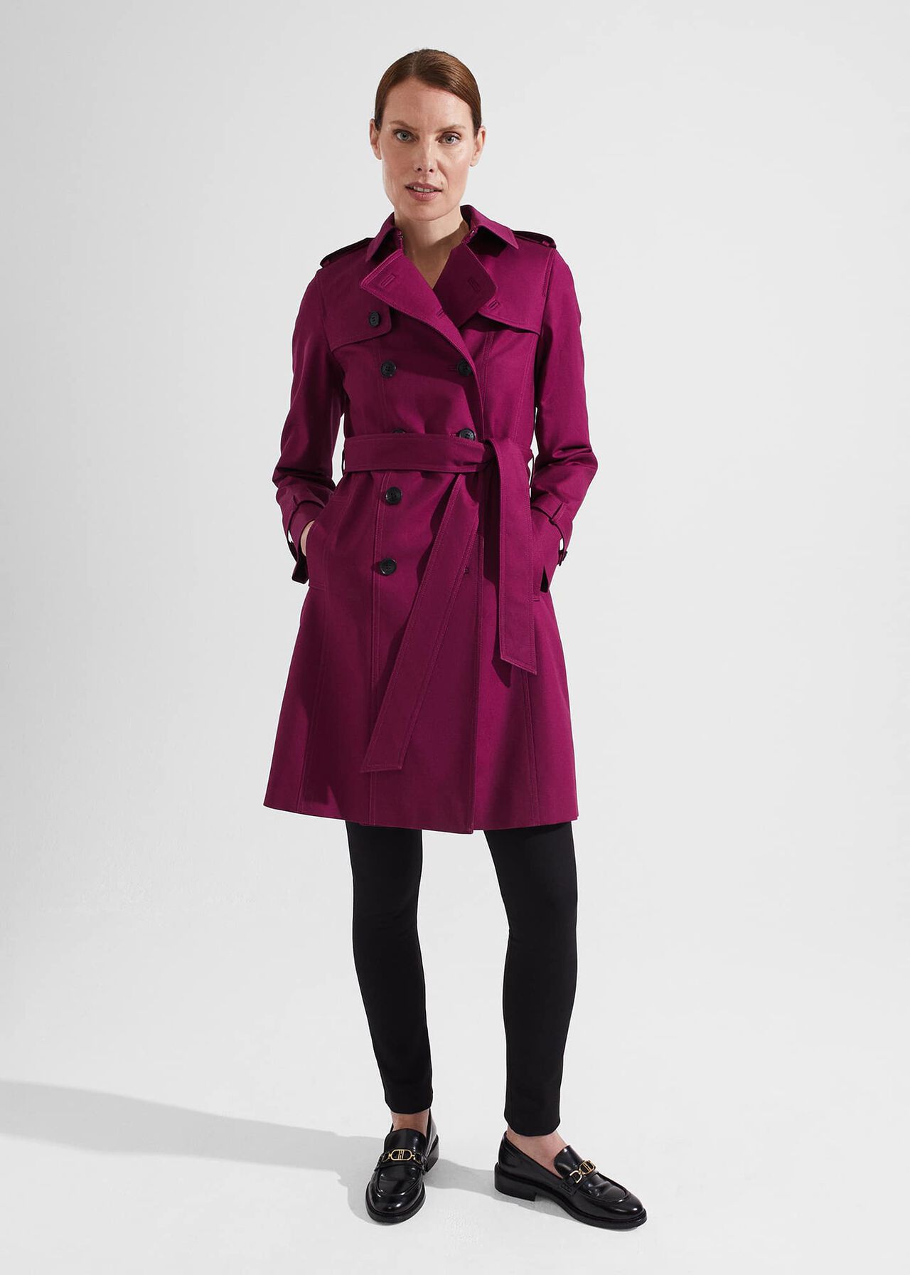 A woman wearing a vibrant fuchsia pink petite trench coat with double-breasted buttons, a waist belt, epaulettes, storm flaps, and a feminine fit and flare silhouette.