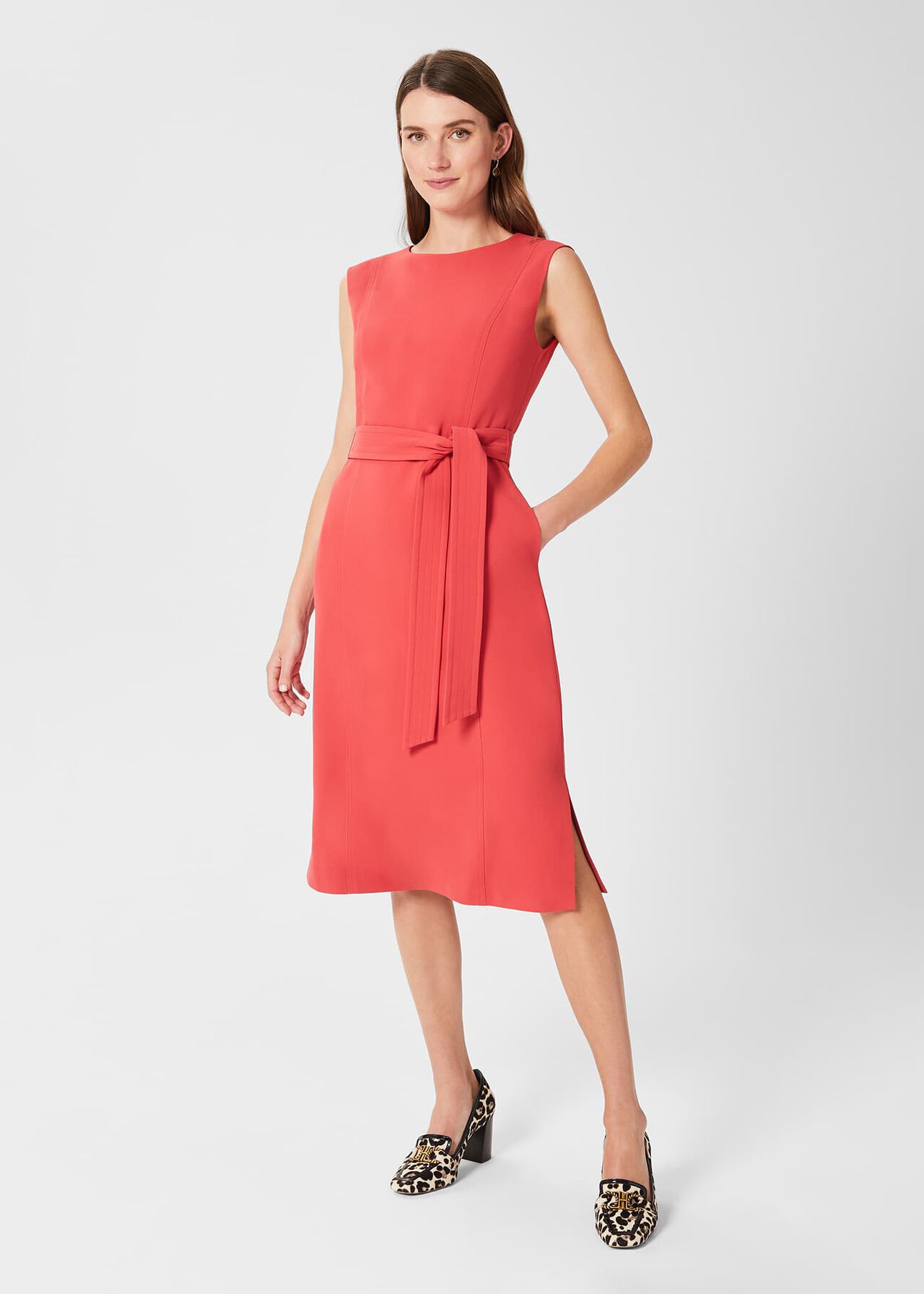A woman wearing a coral sleeveless belted dress with a shift shape, featuring a detachable tie belt and flattering panelling, made from recycled polyester.