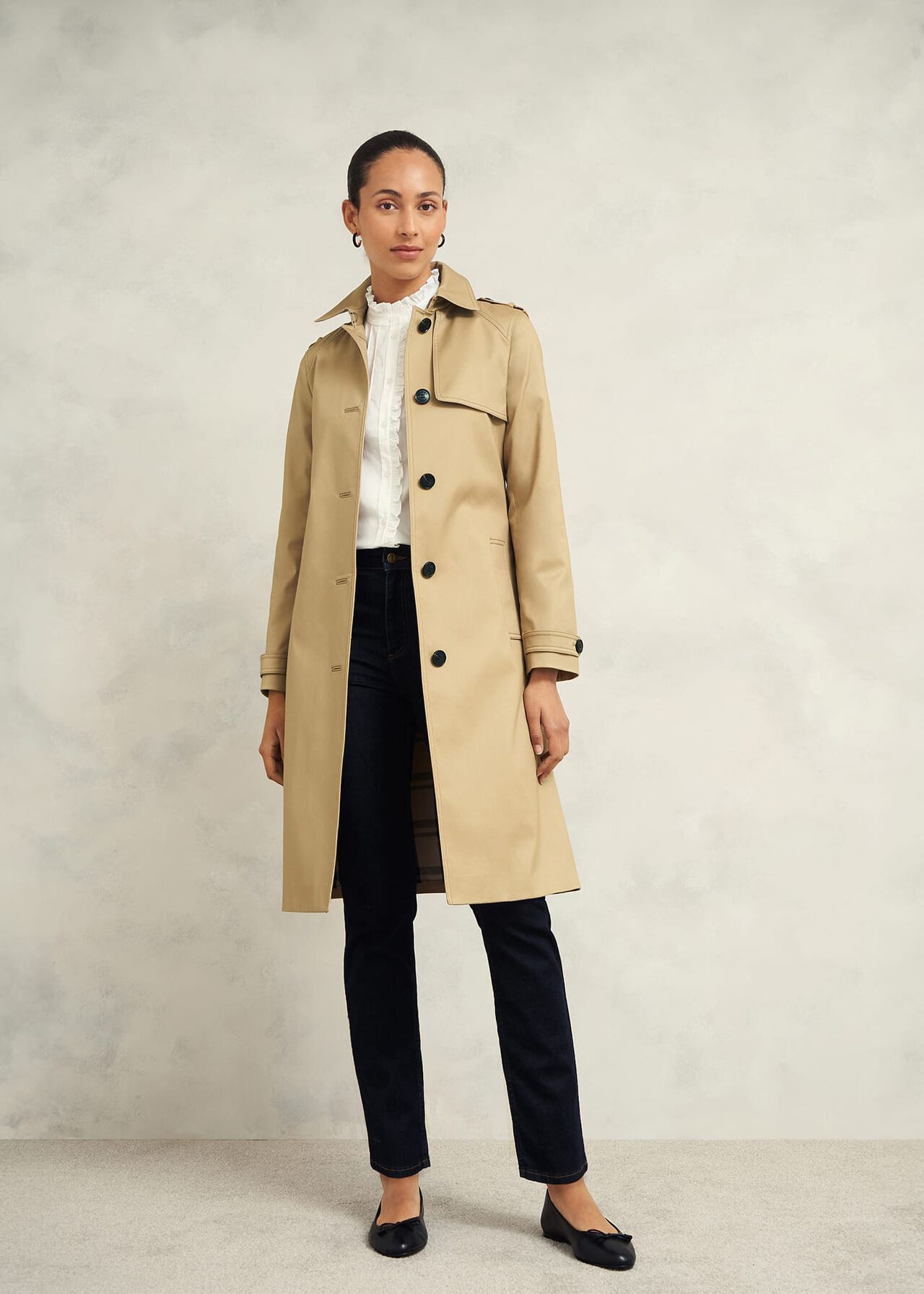 A woman wearing a beige trench coat with a collar, button closure, and weather-ready details, standing against a plain background.
