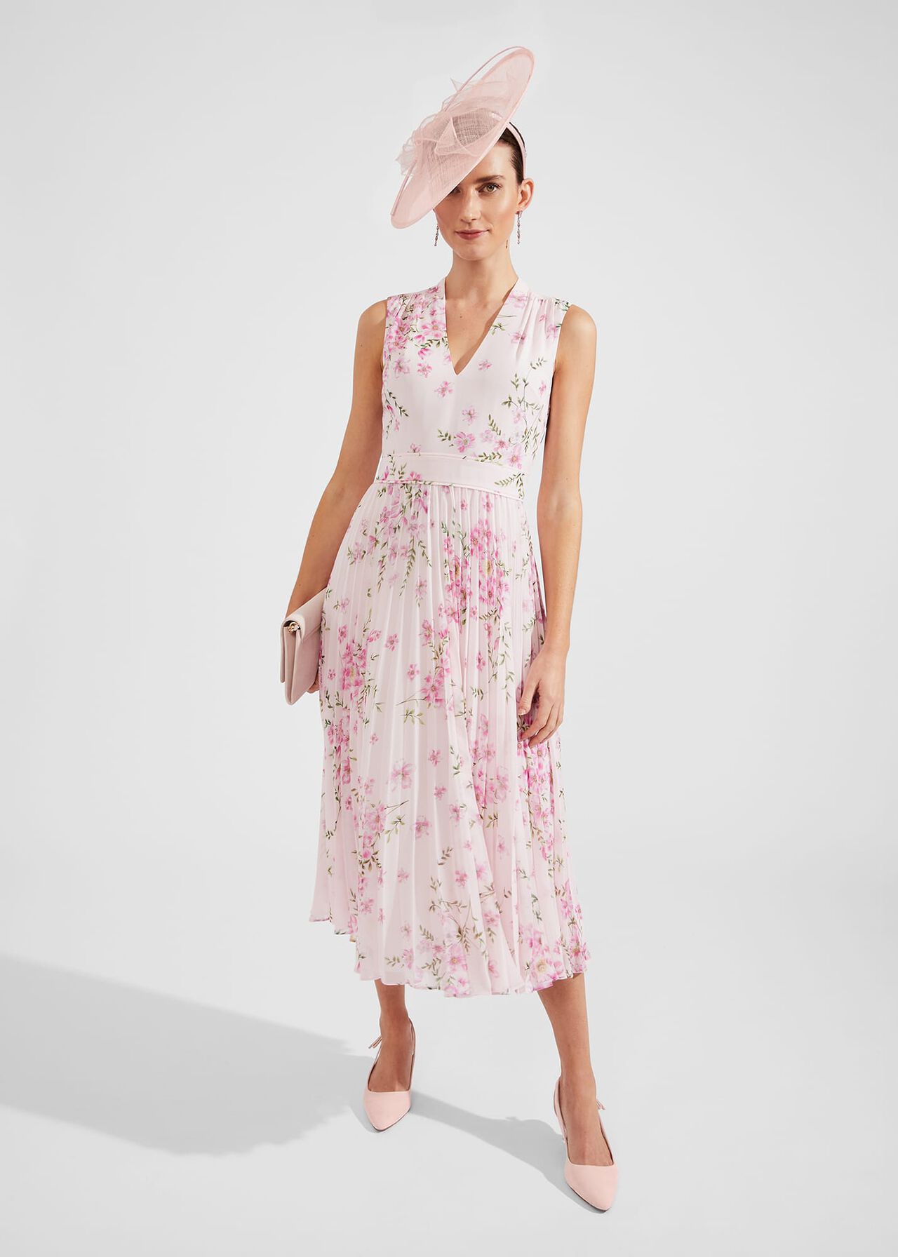 A woman wearing a pink floral pleated dress with a V-neckline, flowing hemline, and floral print, accessorized with a pink hat, clutch, and heels.