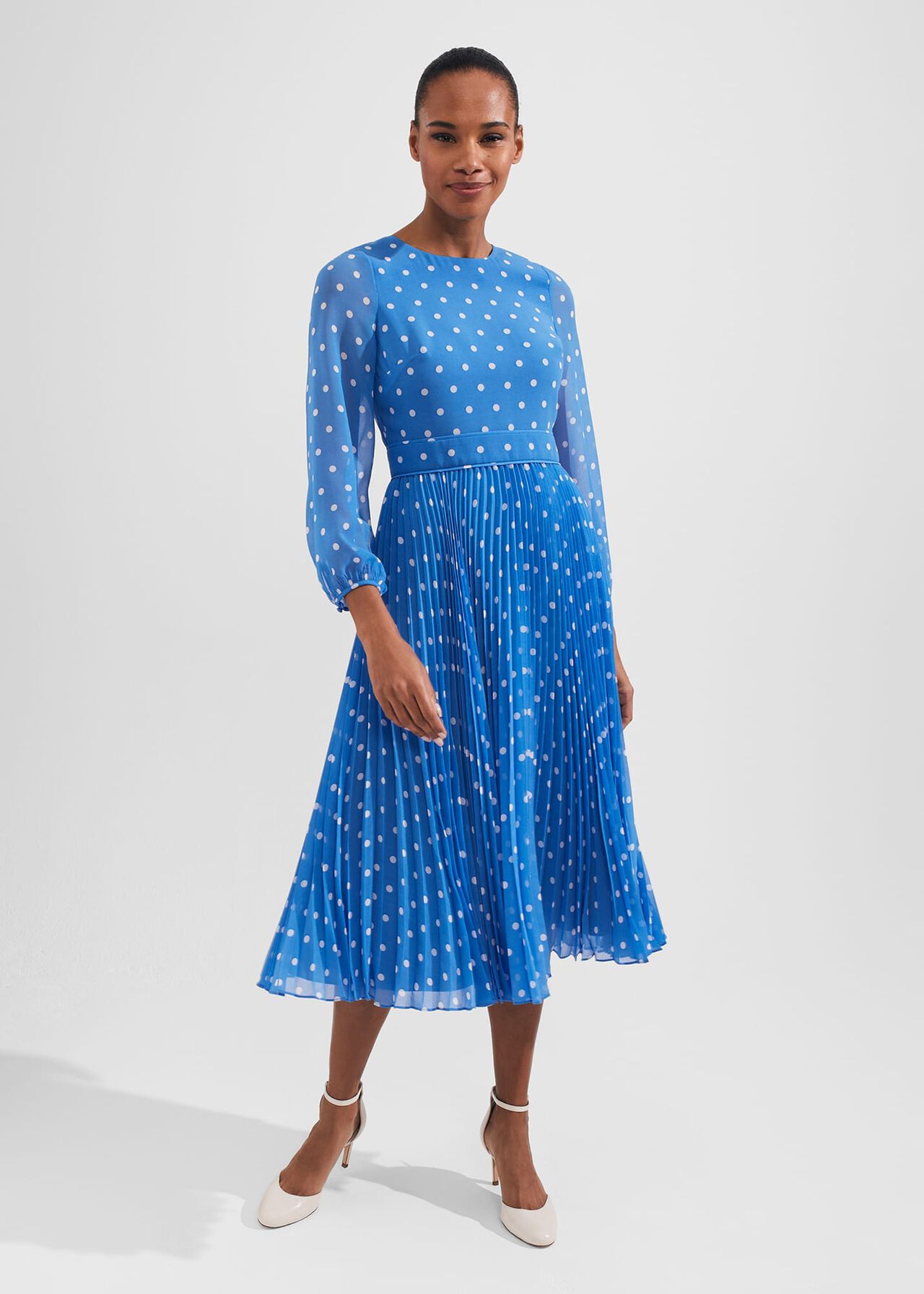A woman wearing a blue fit and flare dress with white polka dots, long puff sleeves, and a pleated skirt.