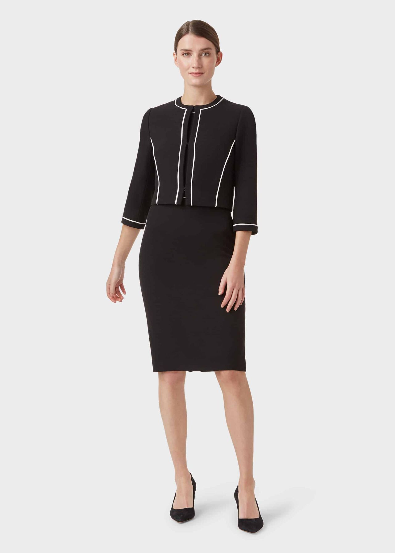 A woman wearing a black, slightly cropped jacket with white piping, round neckline, and three-quarter-length sleeves, paired with a black skirt and black heels.
