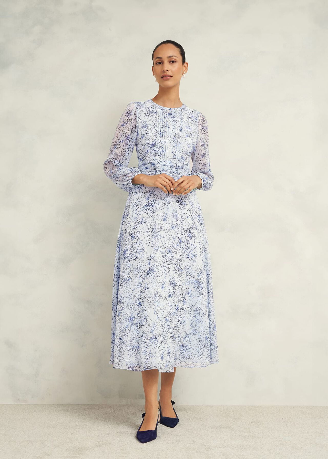 A woman wearing a light blue and white patterned midi dress with long blouson sleeves, gathered waist, and pleated panel, paired with navy heels.