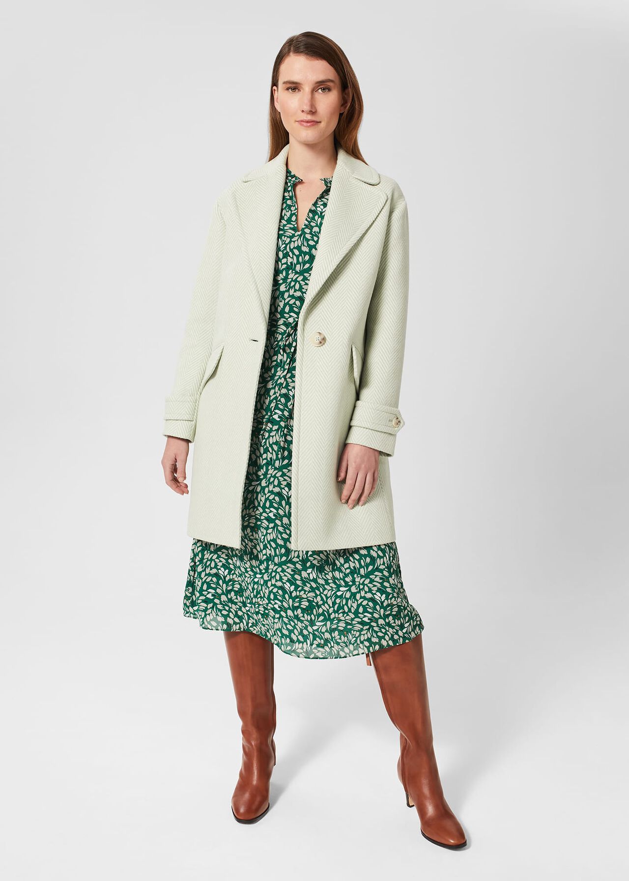 A woman wearing a light beige petite Lillie Coat with a herringbone check pattern, revere collar, and long sleeves, paired with a green patterned dress and brown knee-high boots.