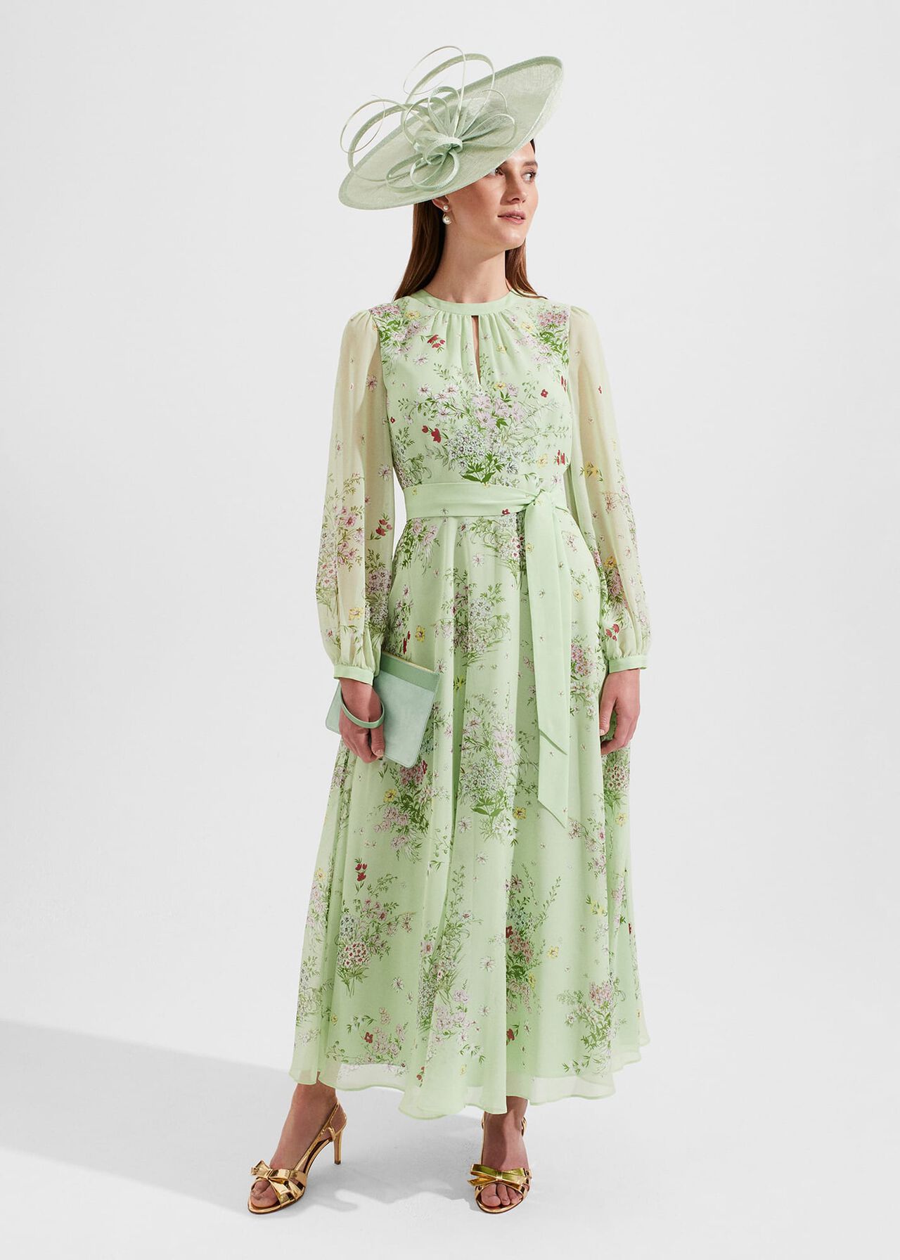 A woman wearing a light green floral silk dress with long sleeves, a tie front, and a flowing hemline, accessorized with a large matching hat and gold heels.
