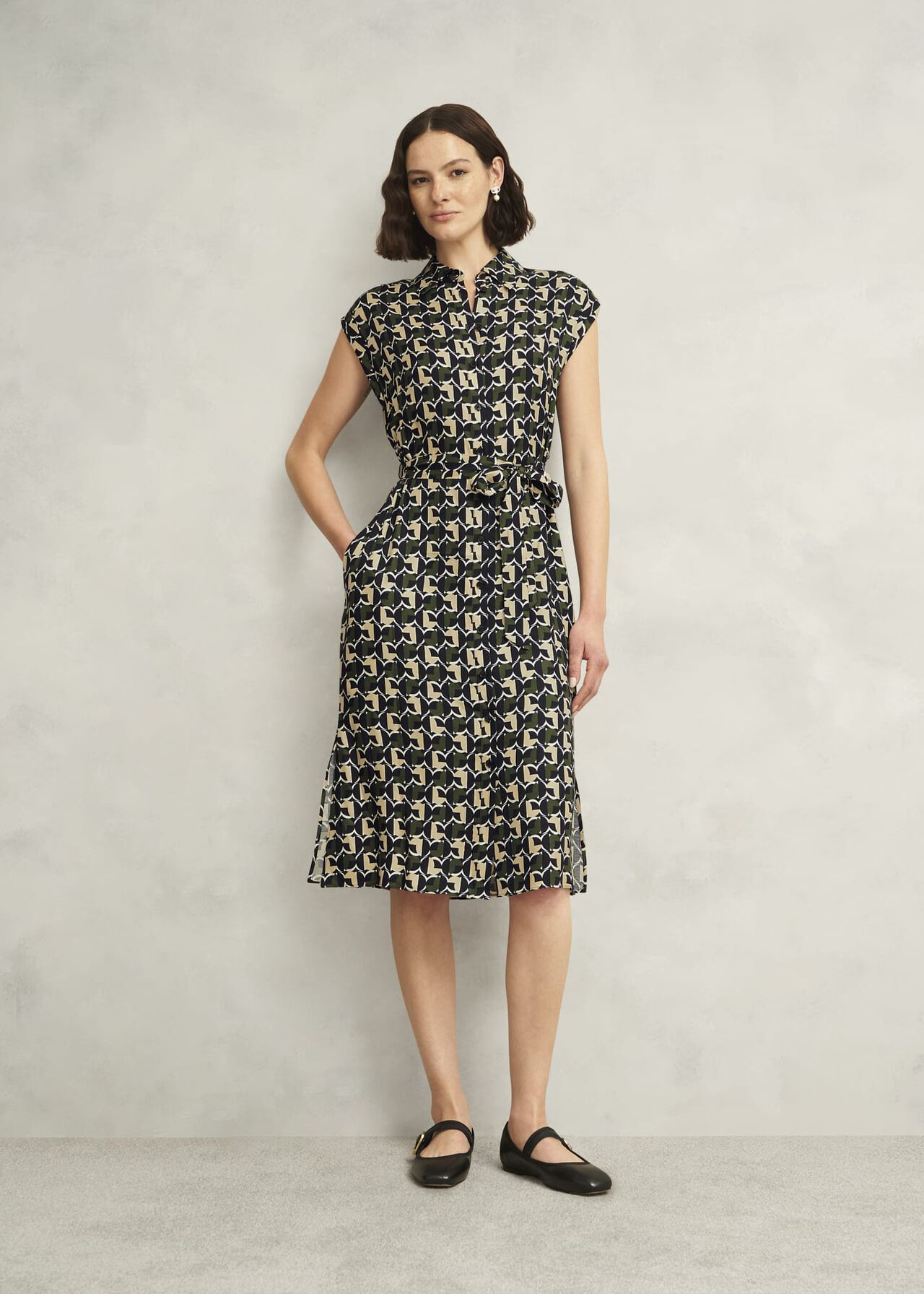 A woman wearing a black, beige, and white geometric print shirt dress with a collar, short sleeves, and a midi length, accessorized with earrings and black shoes.